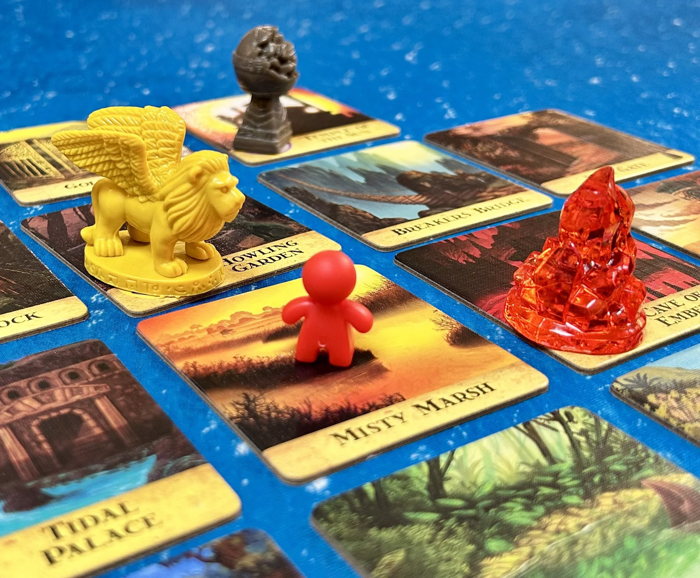 Board game with colorful cards and figurines, including a yellow lion with wings, a red figure, and two crystal-like figures, on a blue surface.