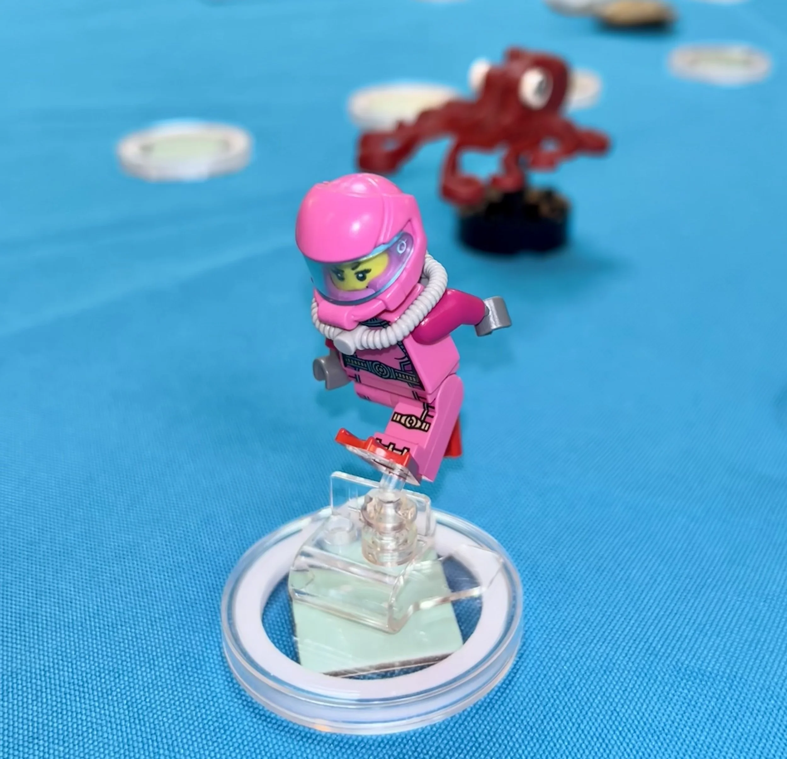 Lego minifigure of a female astronaut in a pink space suit with a helmet, standing on a clear display stand on a blue surface with other blurry Lego figures in the background.