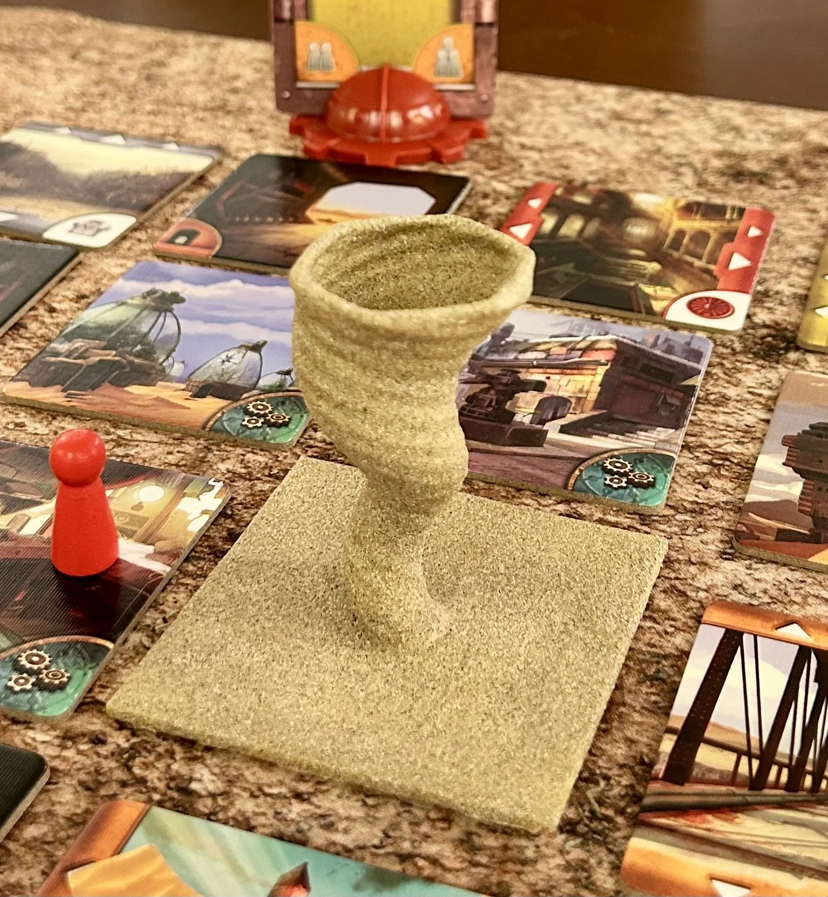 A feedback token shaped like a cone with a spiral design on a textured square base, surrounded by illustrated game cards and a red game piece on a carpeted surface.