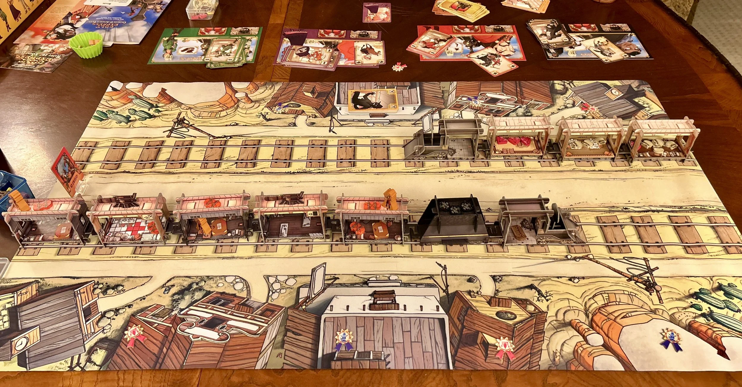 A tabletop board game setup featuring a detailed Western town scene with train tracks, train cars, and miniature buildings, surrounded by various game components and cards.