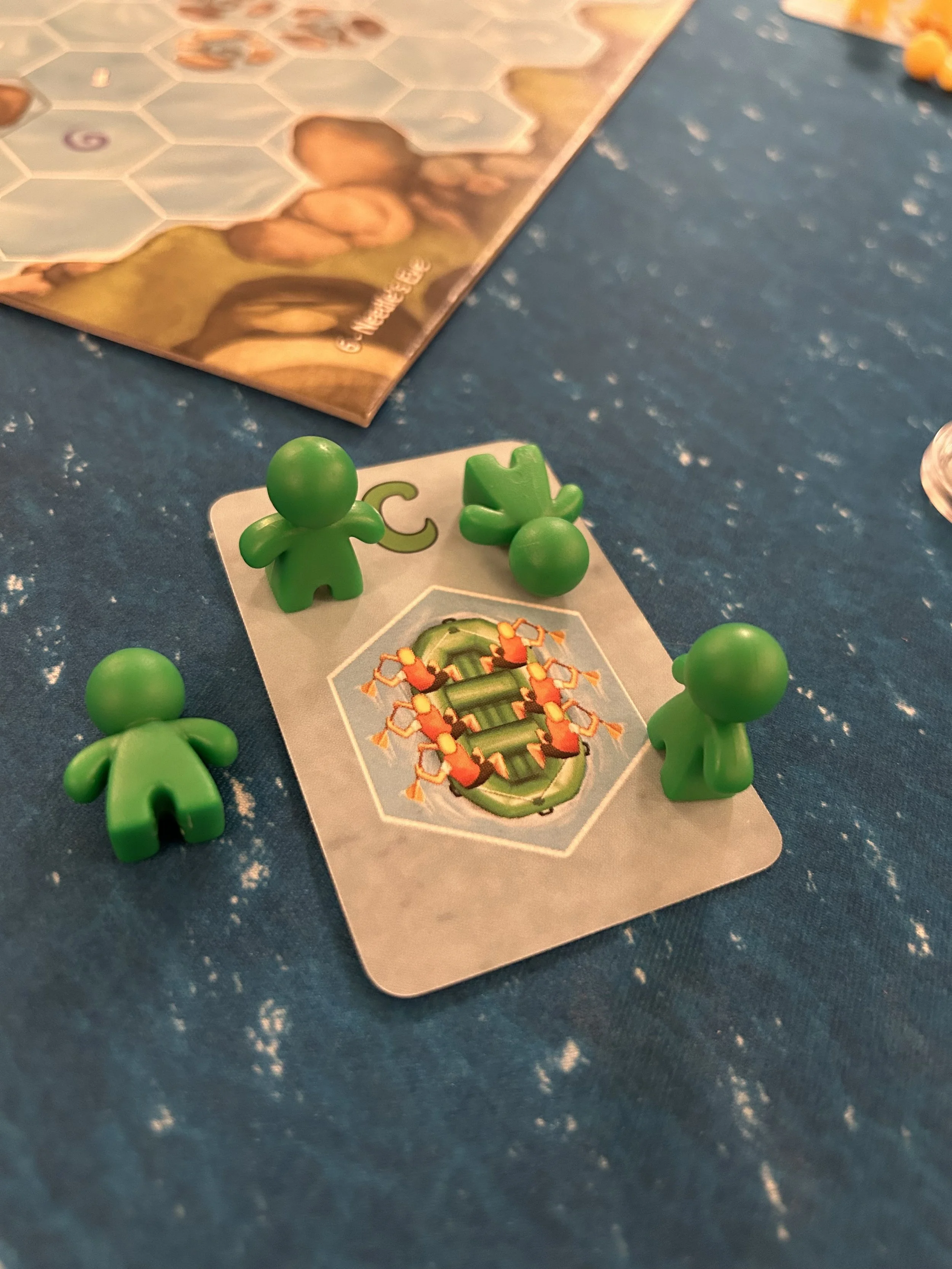 A game piece with four green game pieces, one card with a green logo, and a game board with hexagonal tiles on a blue, speckled surface.