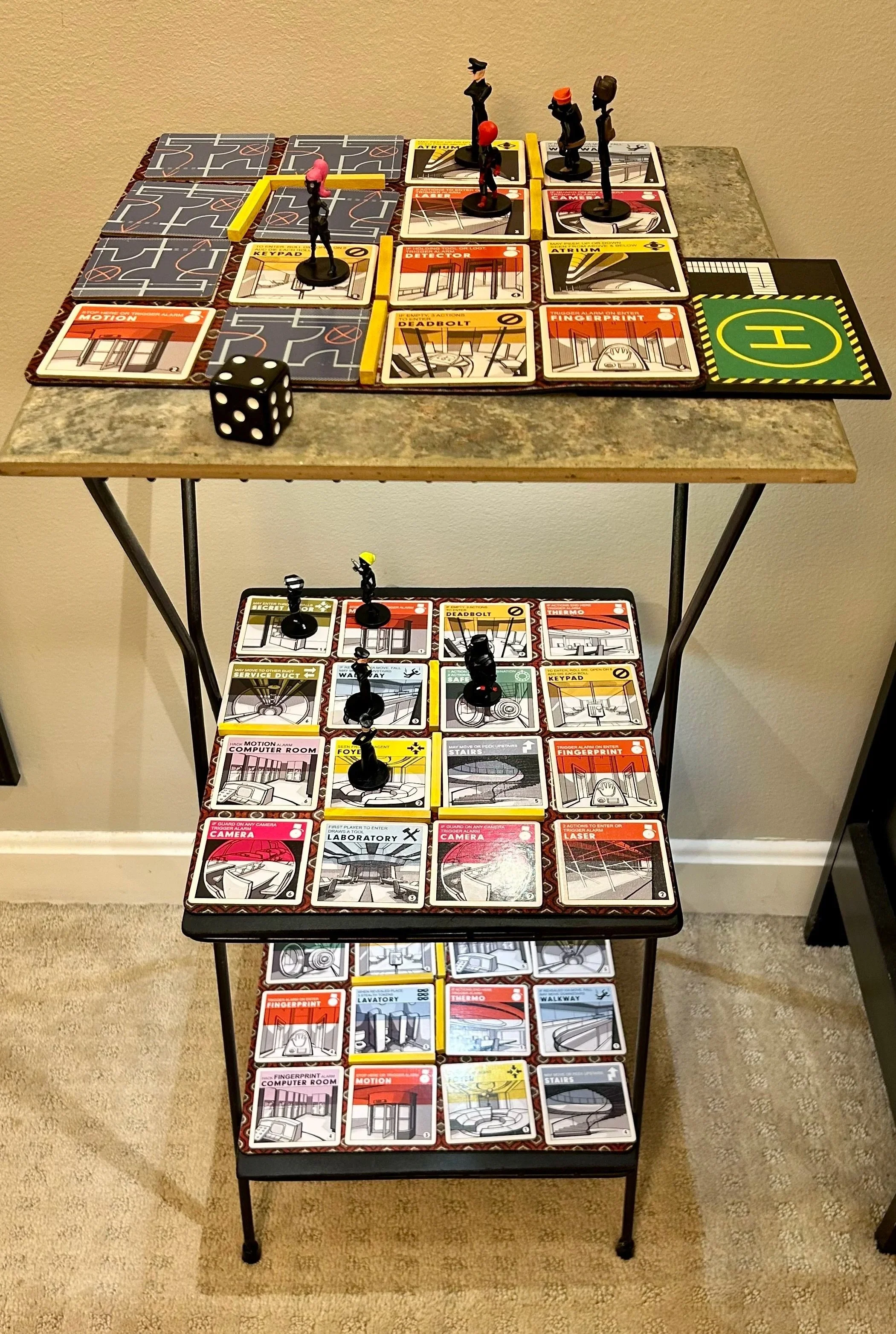Tabletop board game setup with multiple tiles, miniatures, a large black and green helicopter pad, and a black die with white dots.