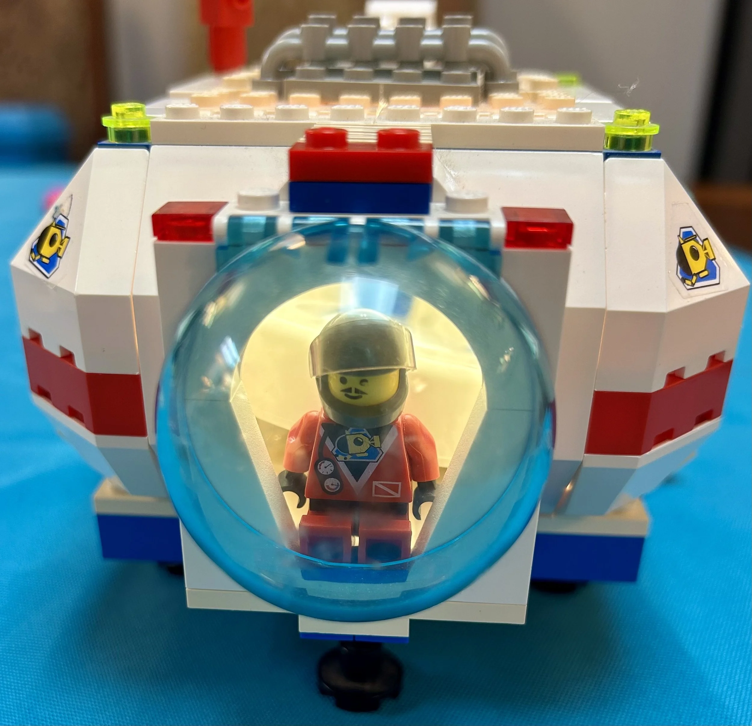 A LEGO space shuttle with a LEGO astronaut minifigure inside, wearing a helmet and a space suit with a camera printed on the torso, front view on a blue surface.