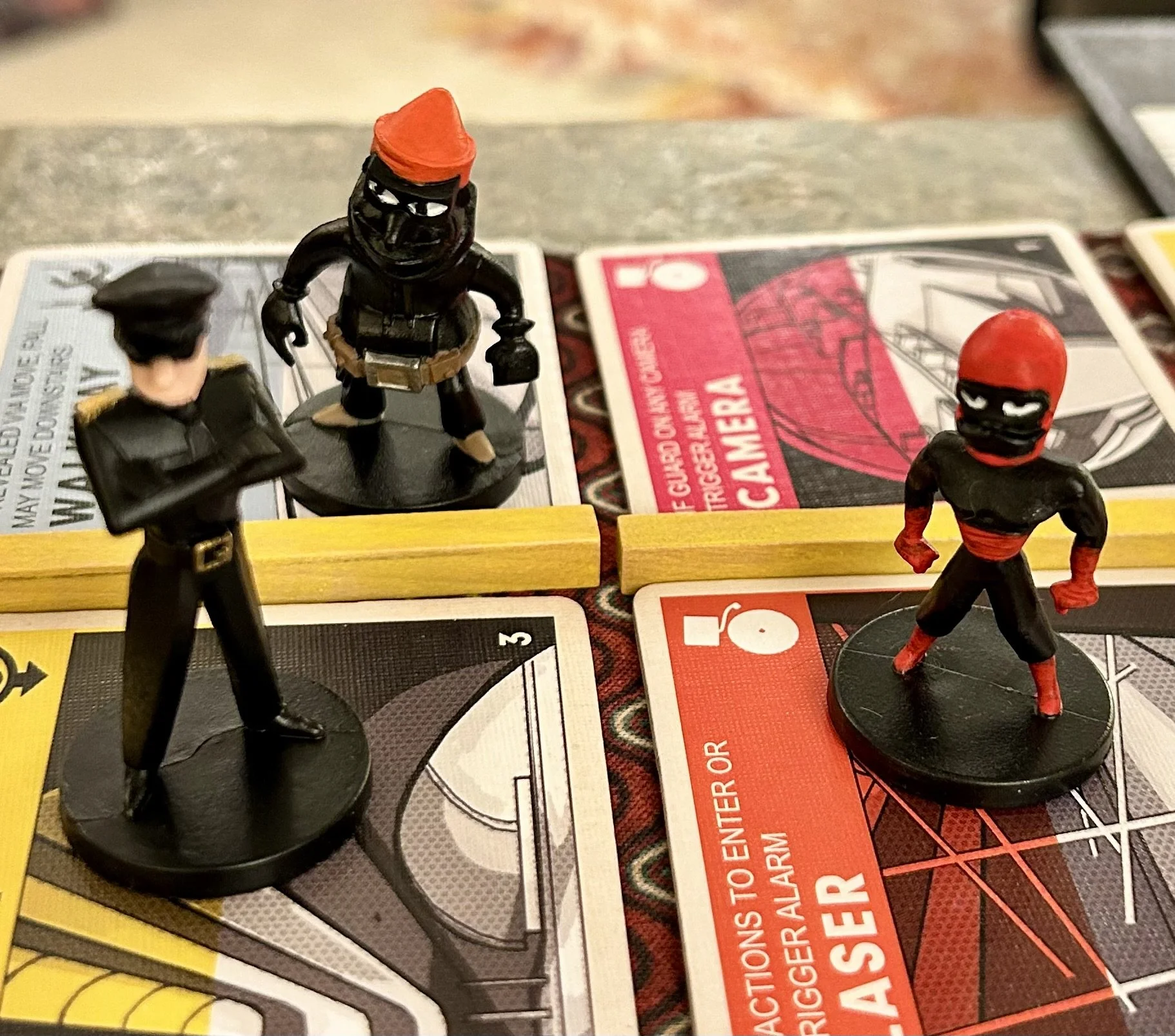 Three small figurines on a board game: a police officer, a black ninja with a red head covering, and a masked ninja with a red head covering and red belt, placed on game cards and yellow blocks.