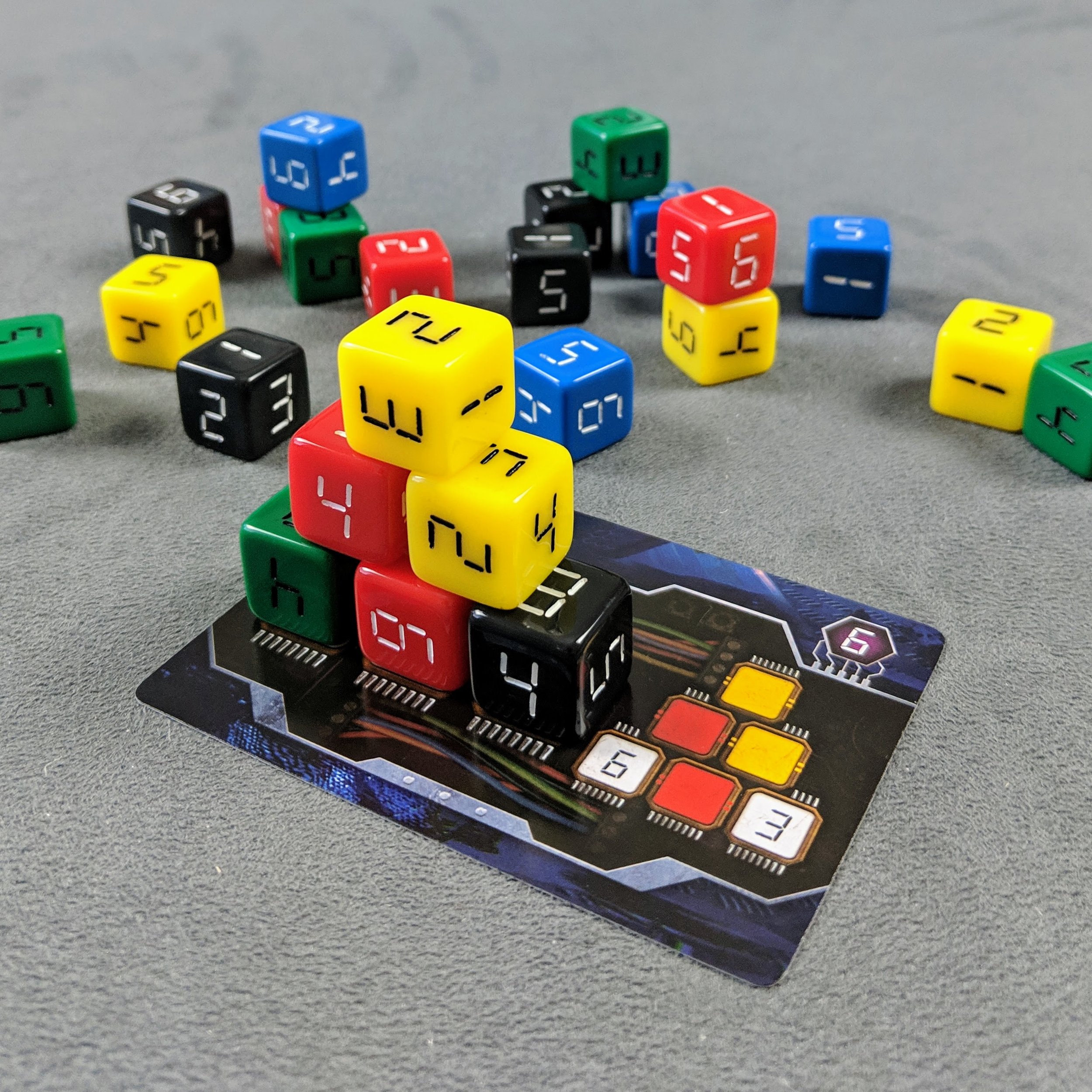 A collection of colorful dice, including yellow, black, red, green, and blue, placed on a gaming card with a futuristic design. Some dice are stacked on top of each other, and others are scattered on a gray surface.