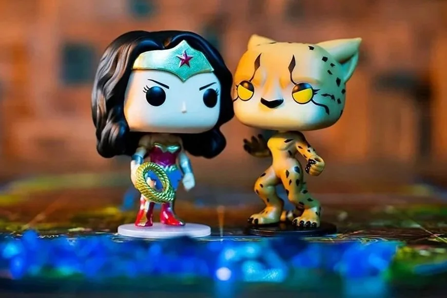 Funko Pop figures of Wonder Woman and a cheetah standing on a game board with a brick wall in the background.