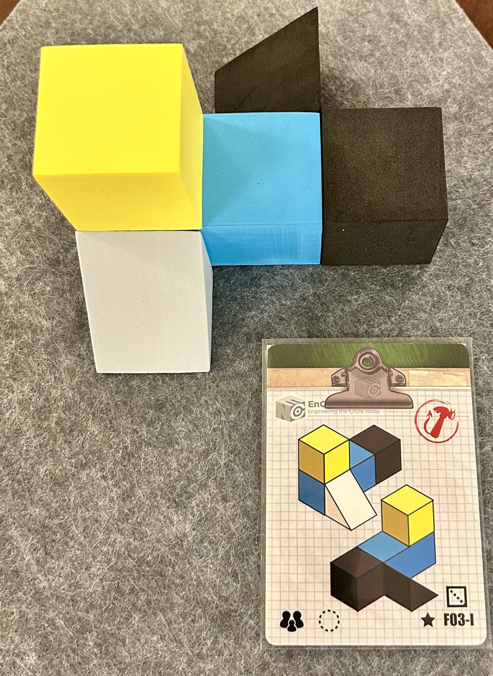 Colorful 3D blocks in yellow, white, blue, and black assembled into a shape, with a game instruction card showing a step-by-step illustration of how to assemble the blocks.