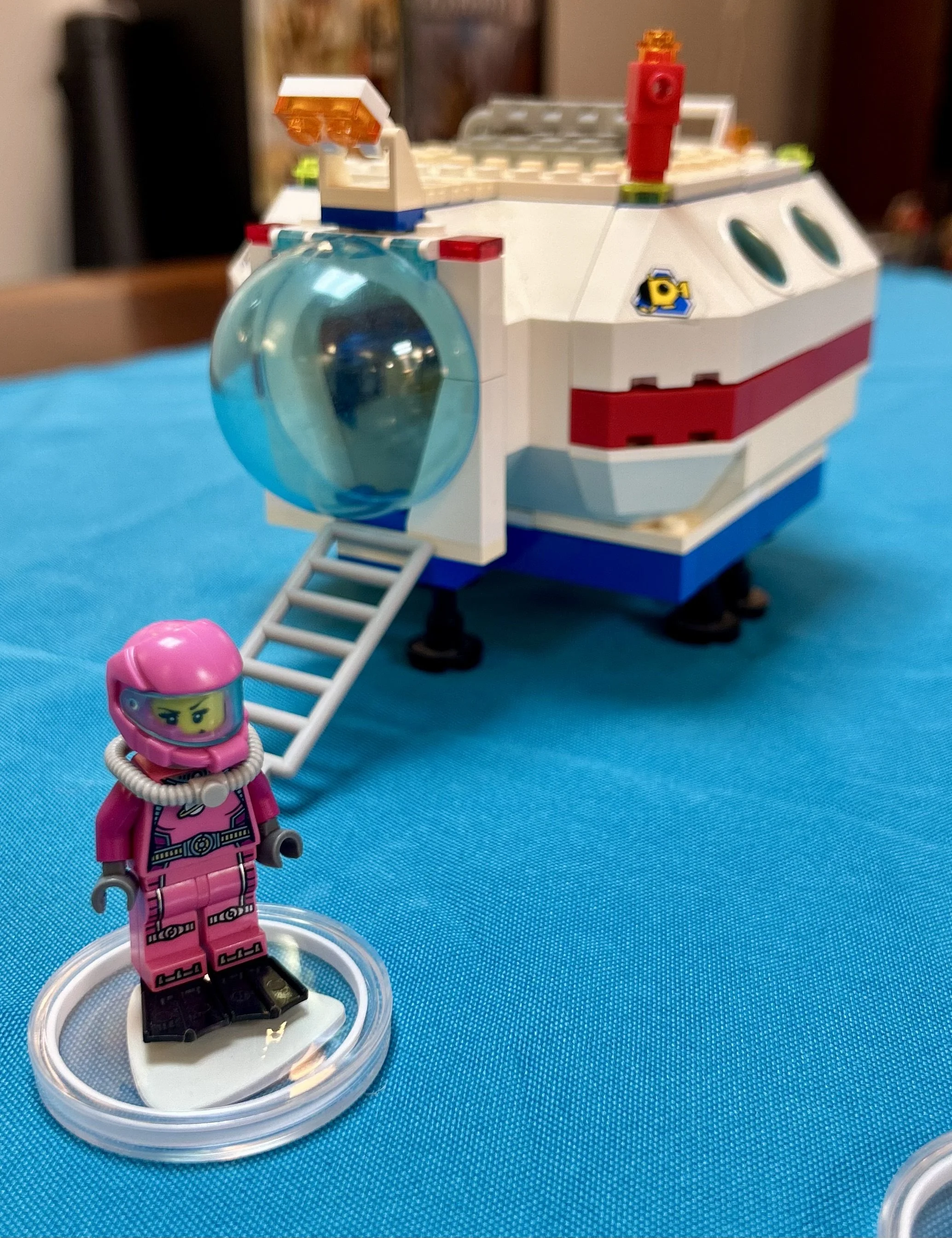 Lego spaceship with a clear spherical cockpit, red, white, and blue body, and a small pink Lego astronaut figurine in a space suit standing on a clear base in the foreground.