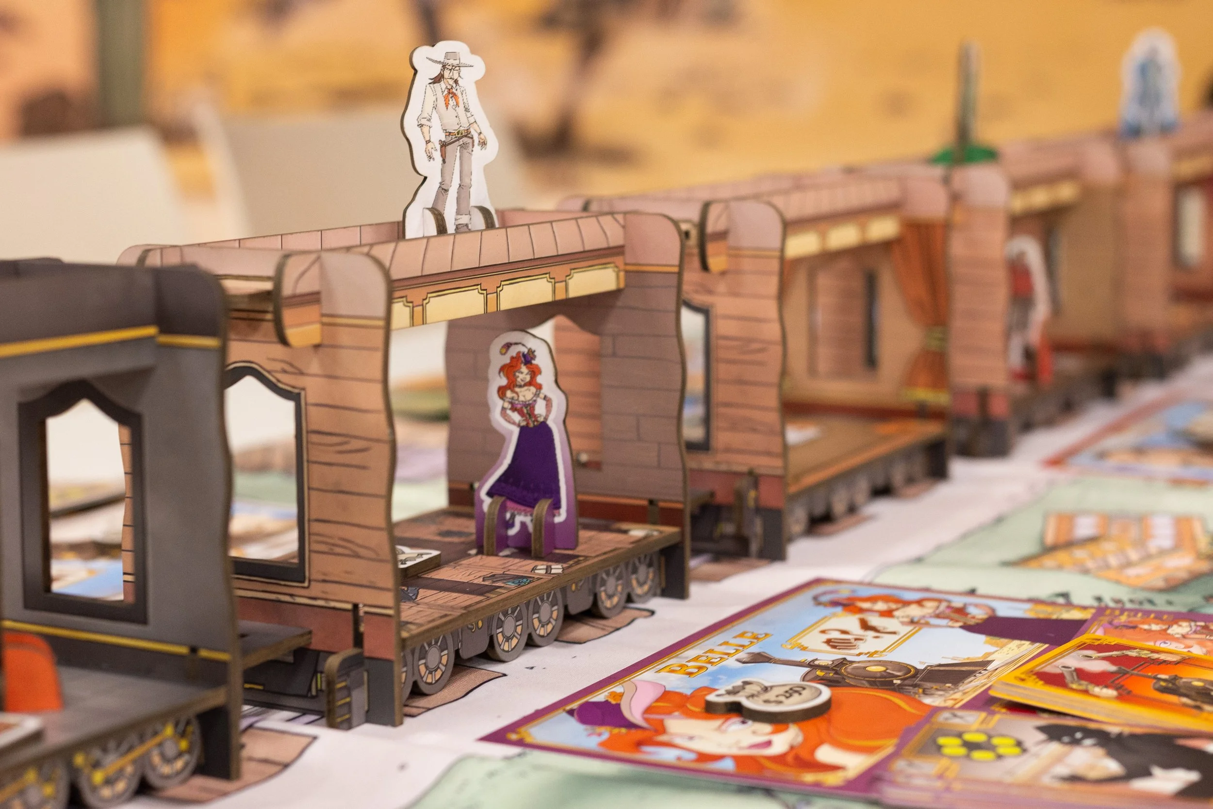 A row of paper-model train cars with cowboy and Western-themed characters on top and colorful game cards and tokens on the table in front.