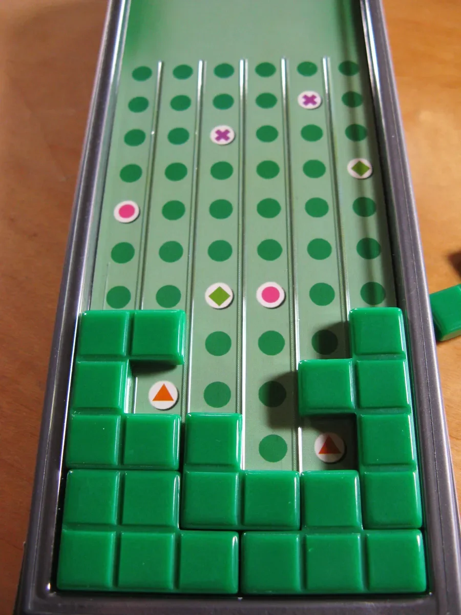 A tabletop game similar to Tetris with green plastic blocks in the lower part of the game board, and colored circles and arrows on the upper game area.