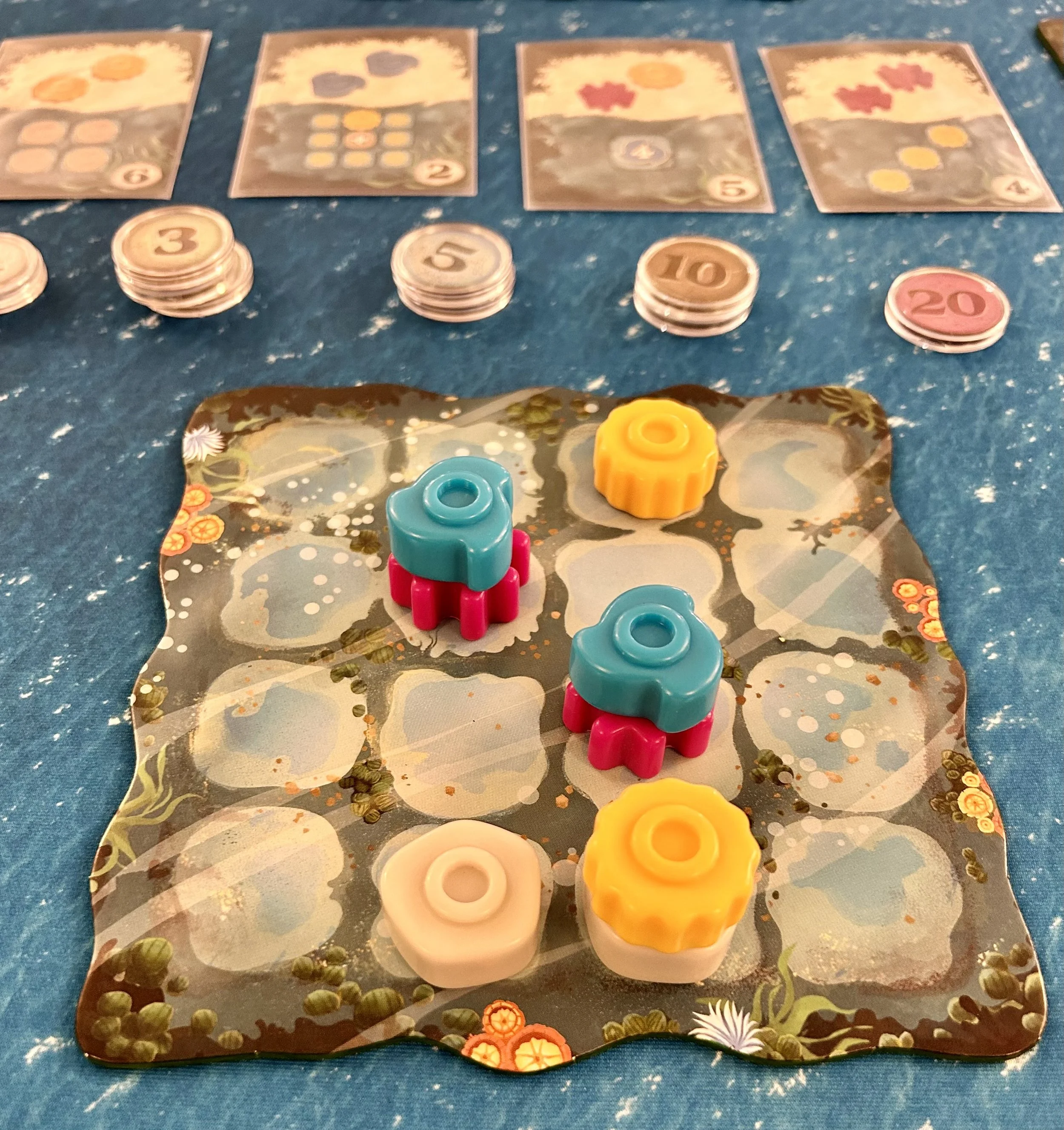 A board game setup with four colorful gear-shaped pieces on a patterned game board, surrounded by game cards and tokens with numbers.