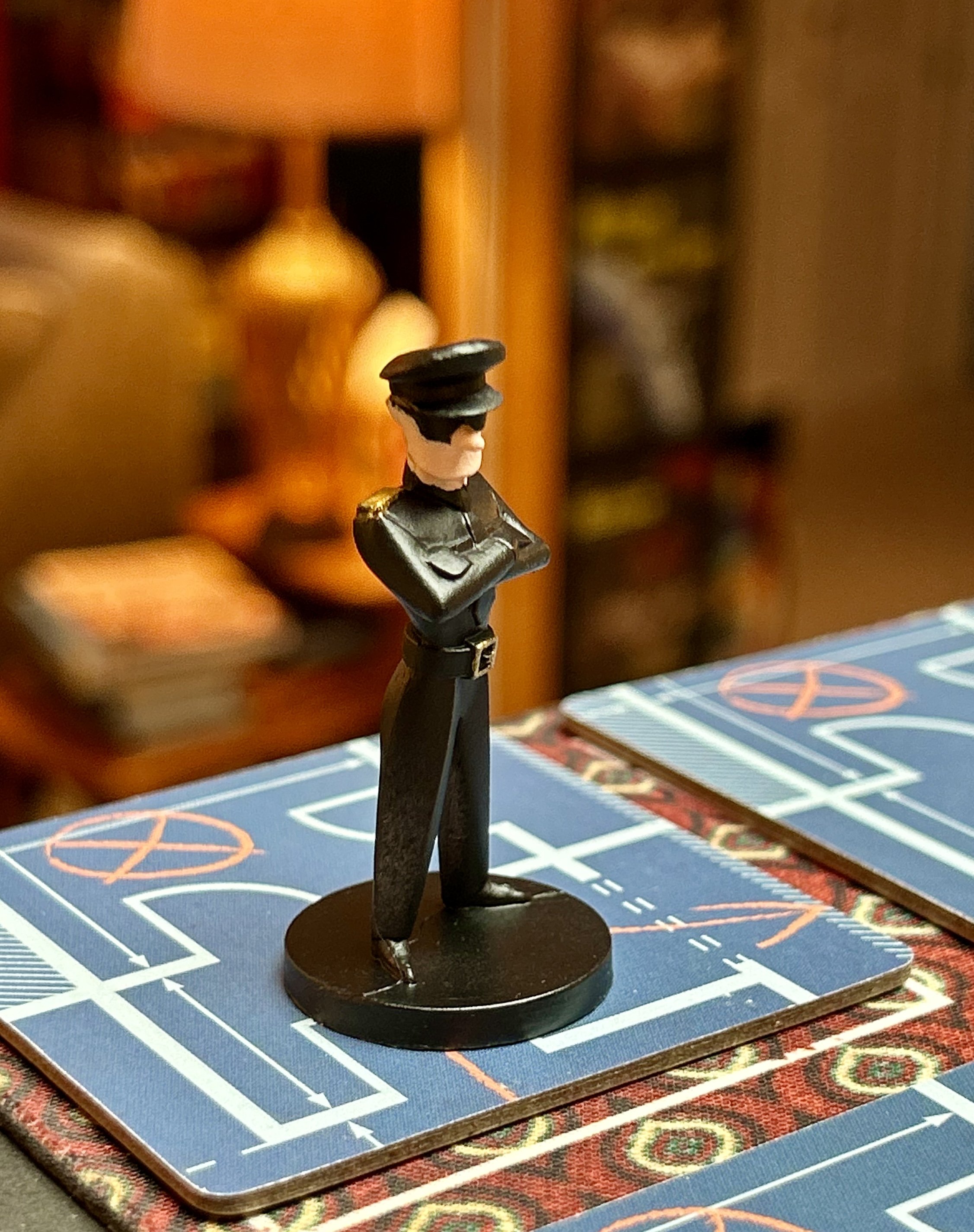 A black figurine of a person dressed as a police officer, standing with arms crossed on a pieced-together game board with blue, white, and red designs.