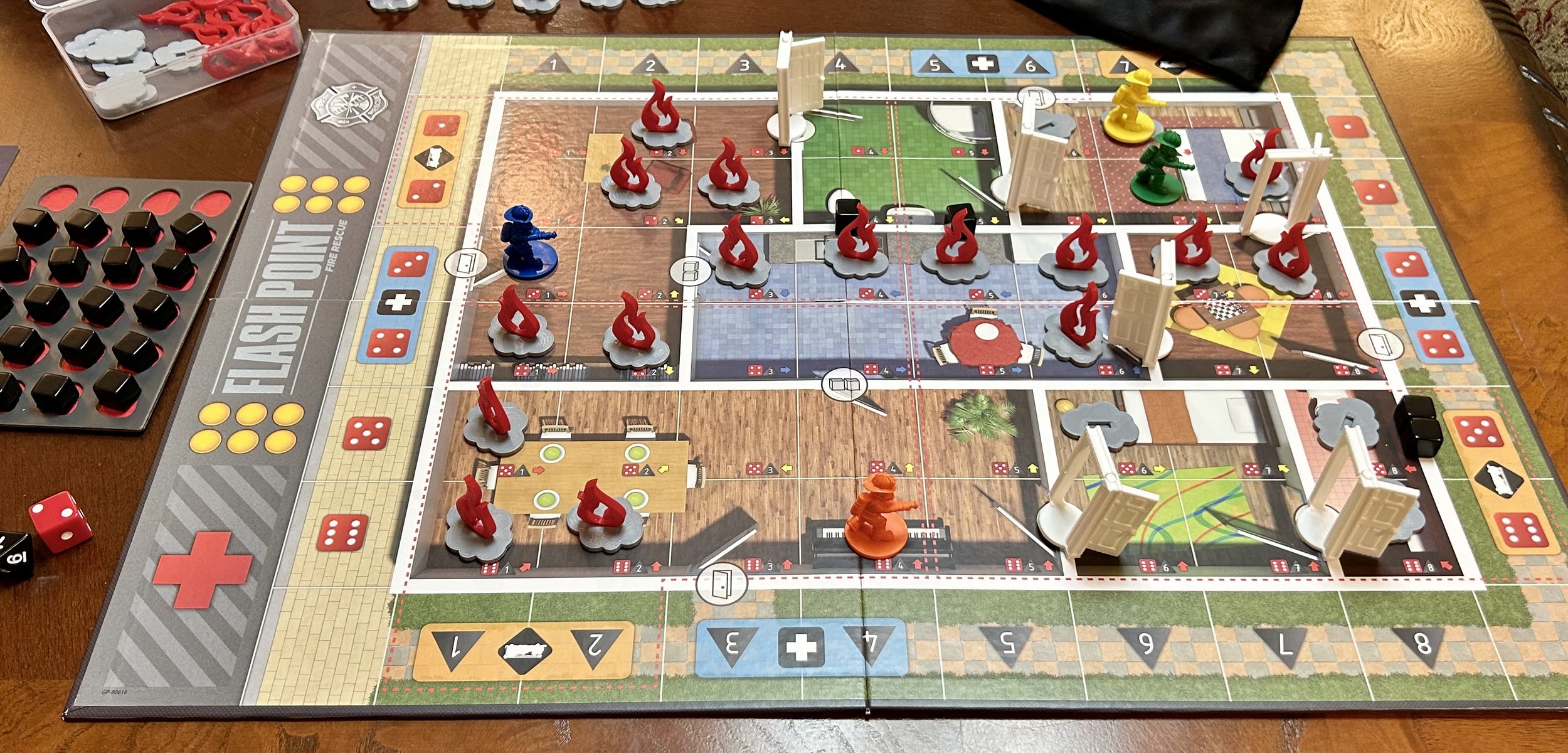 A colorful board game with various game pieces, including miniature figures, dice, and cards, set up on a wooden table. The game features different zones with illustrations of rooms and themes, and there are containers holding additional game tokens and accessories.