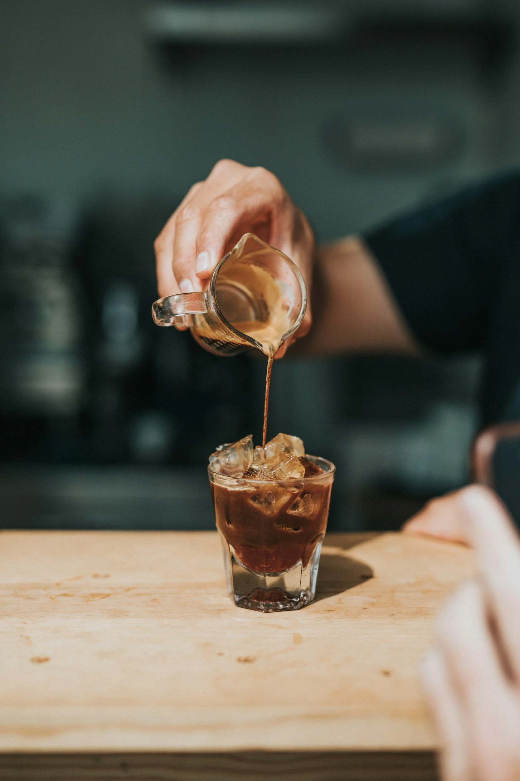 A person pouring coffee or espresso from a glass pitcher into a glass filled with ice on a wooden surface.