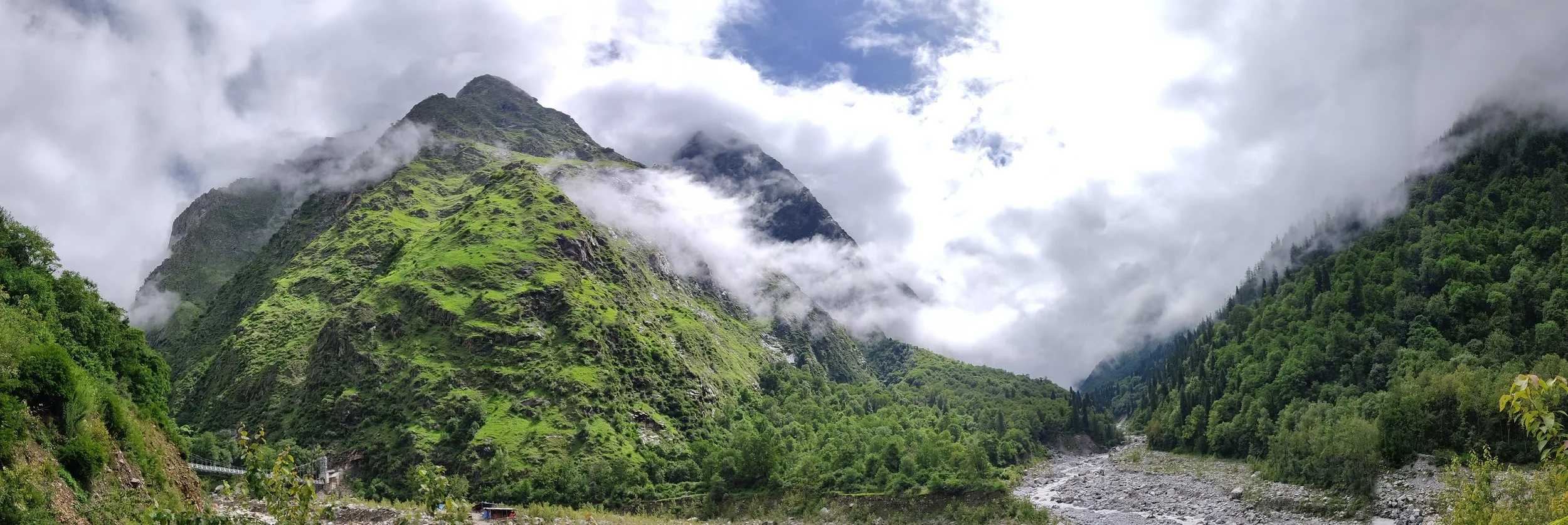 Green mountain landscape with clouds and fog, a river flowing through the valley, lush trees on the hillside.