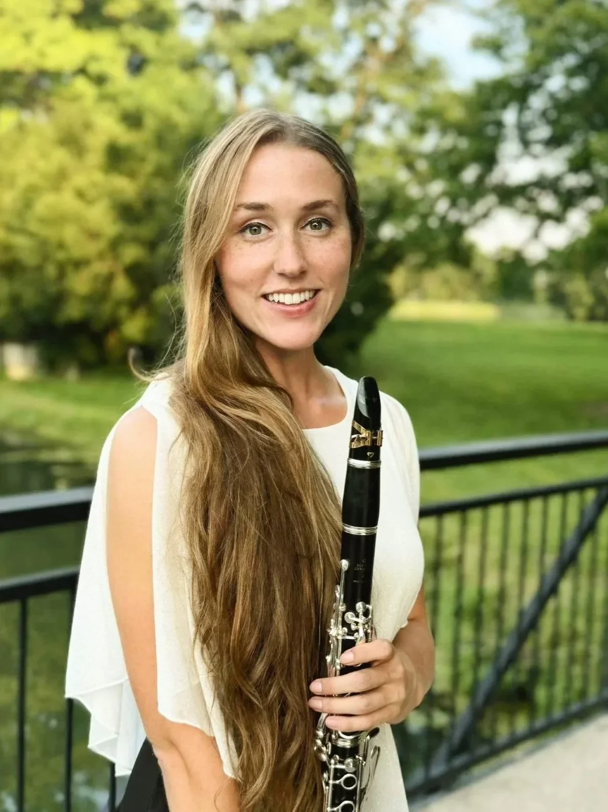 A smiling young woman with long, wavy blonde hair poses outdoors holding a clarinet, with greenery and trees in the background.