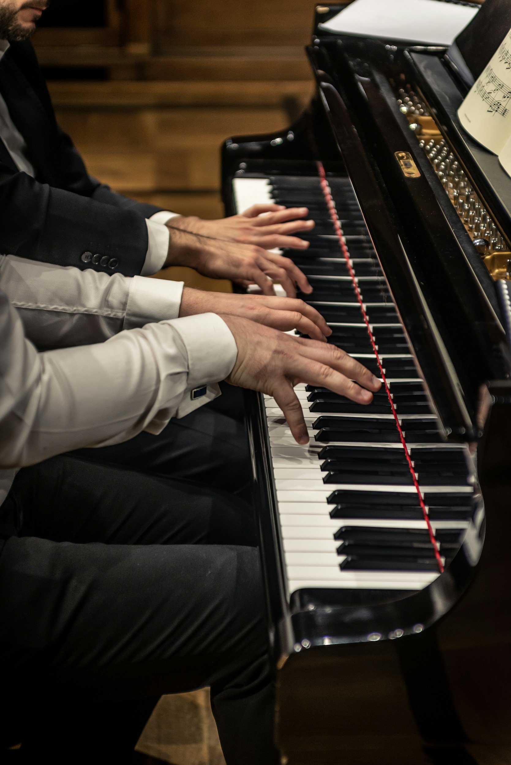 Two people playing a grand piano together, with sheet music on the stand, in a room with wooden walls.