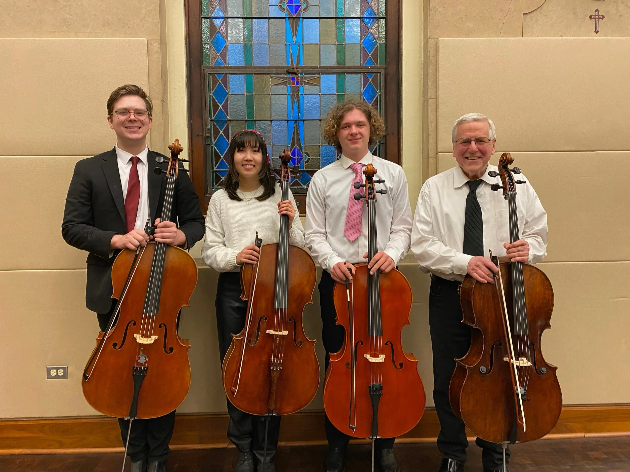 Group of four musicians holding cellos, standing in front of a stained glass window in a church or concert hall.