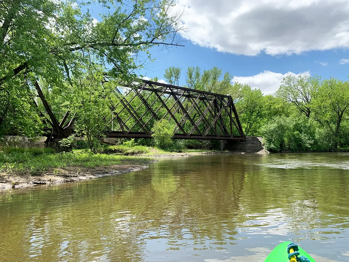 A partially collapsed black metal bridge over a river, surrounded by lush green trees under a partly cloudy sky, with a green kayak visible in the lower right corner.