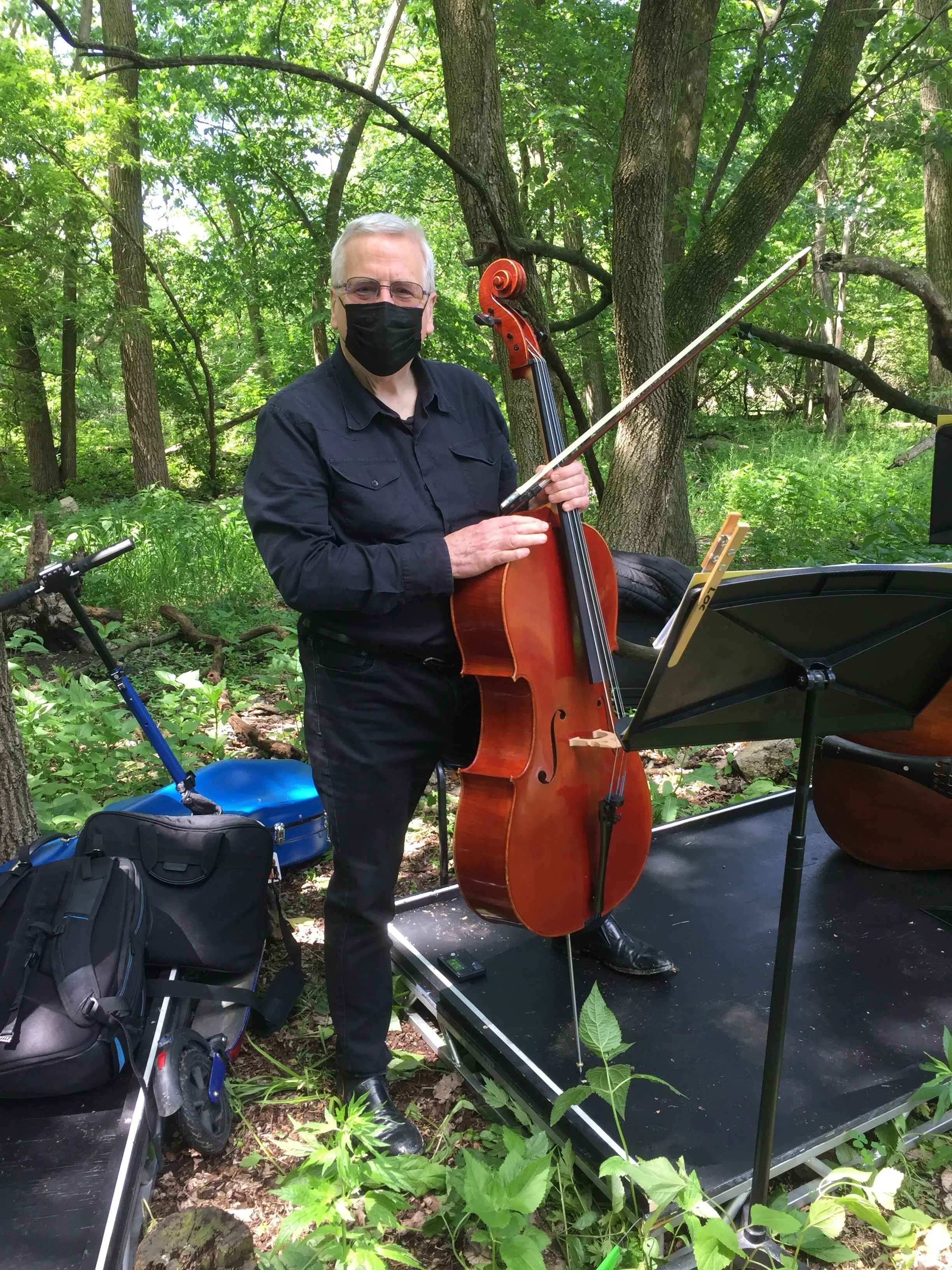 A man wearing a black face mask and black clothing is playing a cello outdoors among trees and green foliage, standing on a small stage with some equipment and a music stand.