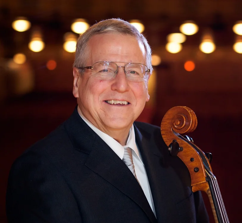 A smiling man wearing glasses, a dark suit, a white shirt, and a tie, holding a violin. The background features blurred warm lights.