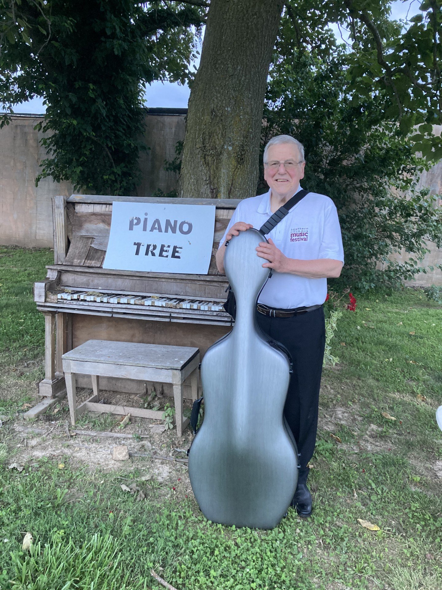 An elderly man in a white shirt and black pants is standing outdoors, holding a large violin case. He is smiling and standing next to a weathered outdoor piano with a sign that reads "PIANO TREE". The background features a large tree, greenery, and a stone wall.