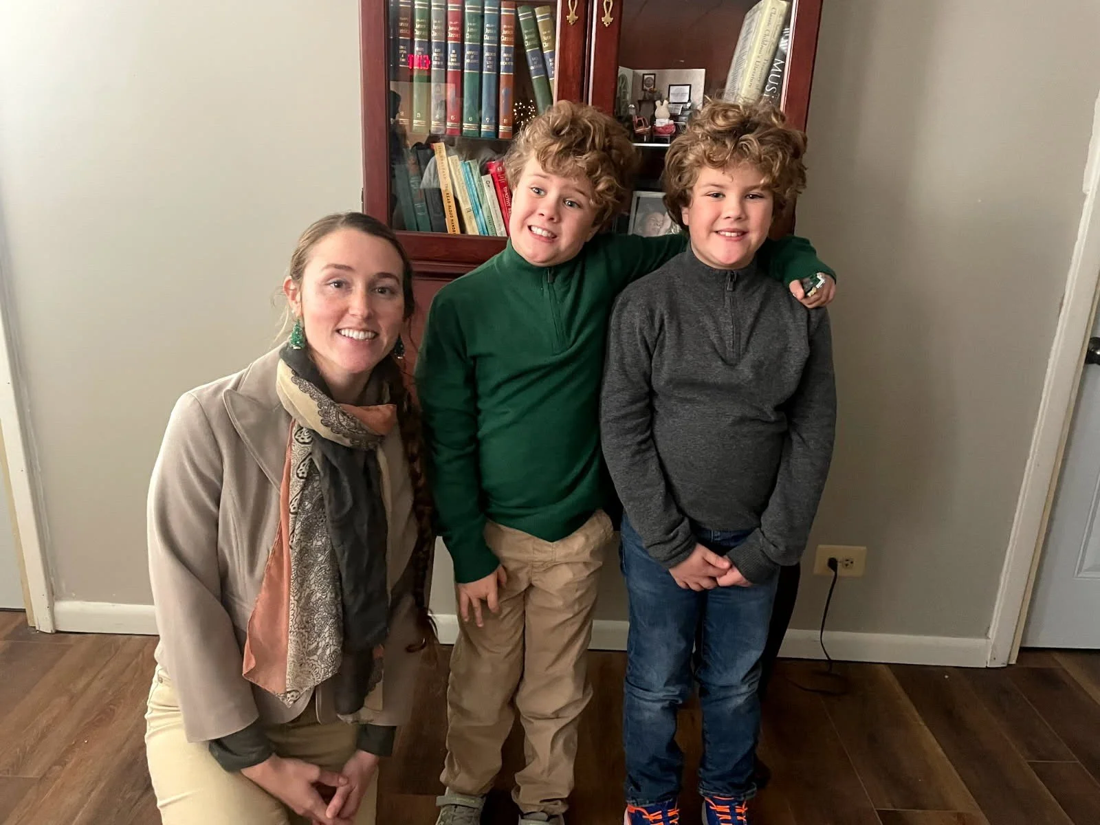 A woman and two boys standing in a room with a bookcase in the background. The woman is kneeling on the left, wearing a beige jacket and scarf, smiling. The boys are standing side by side, both with curly hair and smiling, with one wearing a green shirt and the other a dark gray shirt.