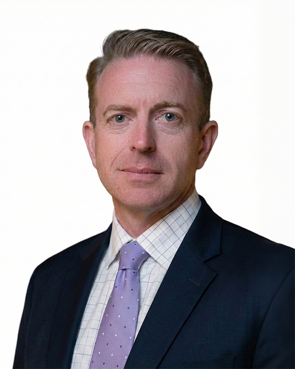A professional headshot of a middle-aged man with short blonde hair, wearing a dark business suit, light checkered shirt, and light purple tie, against a plain white background.