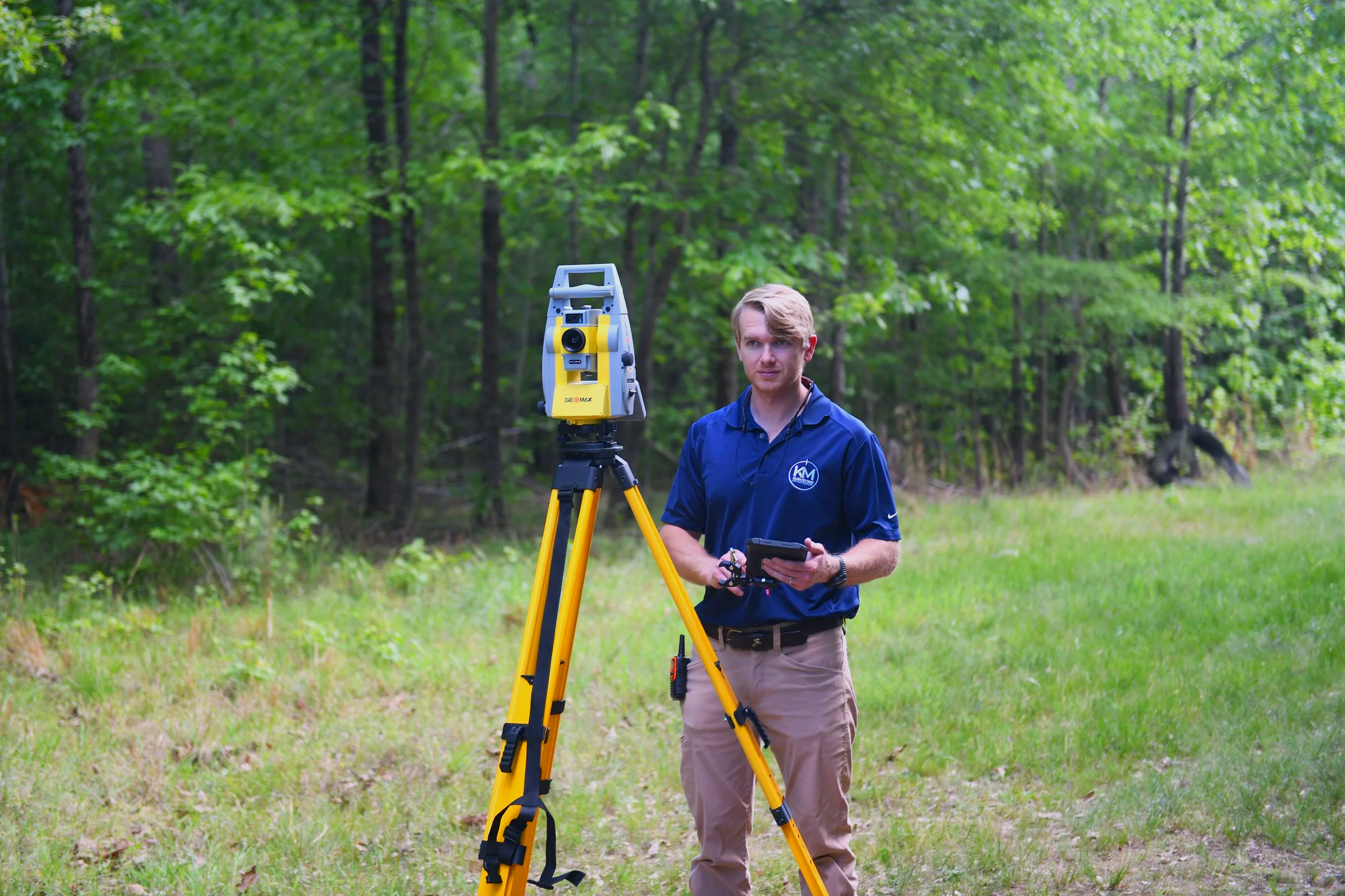 A man in a blue shirt stands outdoors in a forested area operating a yellow and gray surveying instrument on a tripod, holding a remote control or tablet.