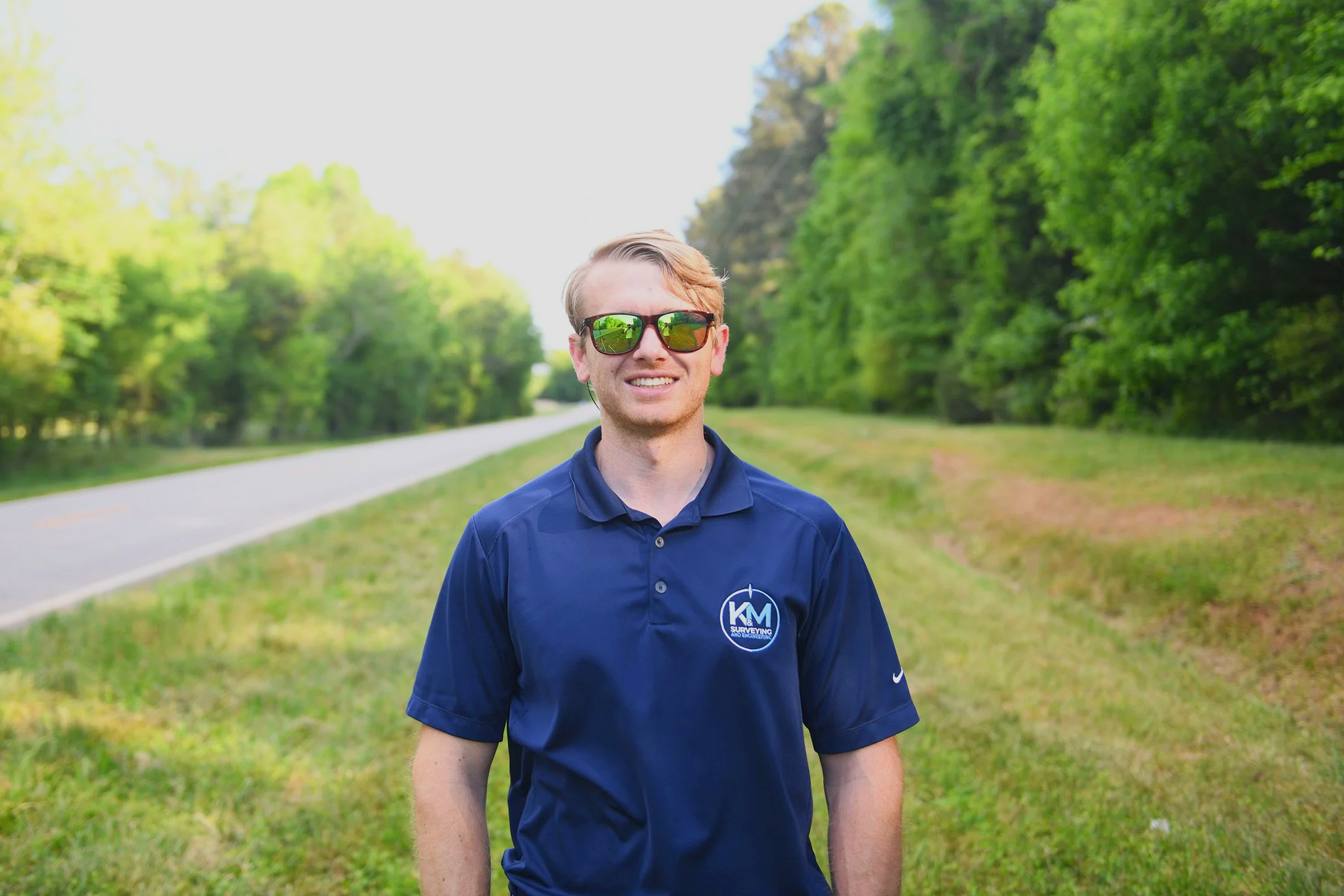 A young man with blonde hair and sunglasses smiling outdoors, wearing a blue polo shirt with a logo, standing on a grassy area near a tree-lined road.