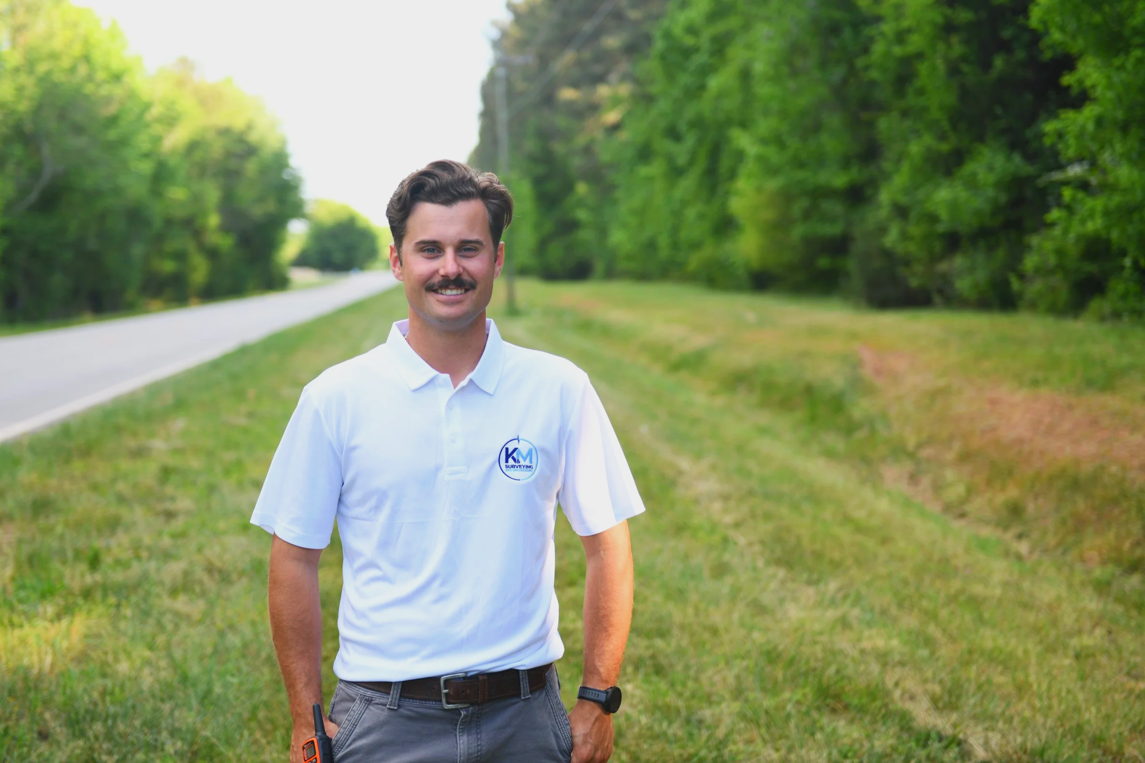 A man dressed in a white shirt with a logo, standing outdoors on a grassy area with trees in the background, smiling at the camera.