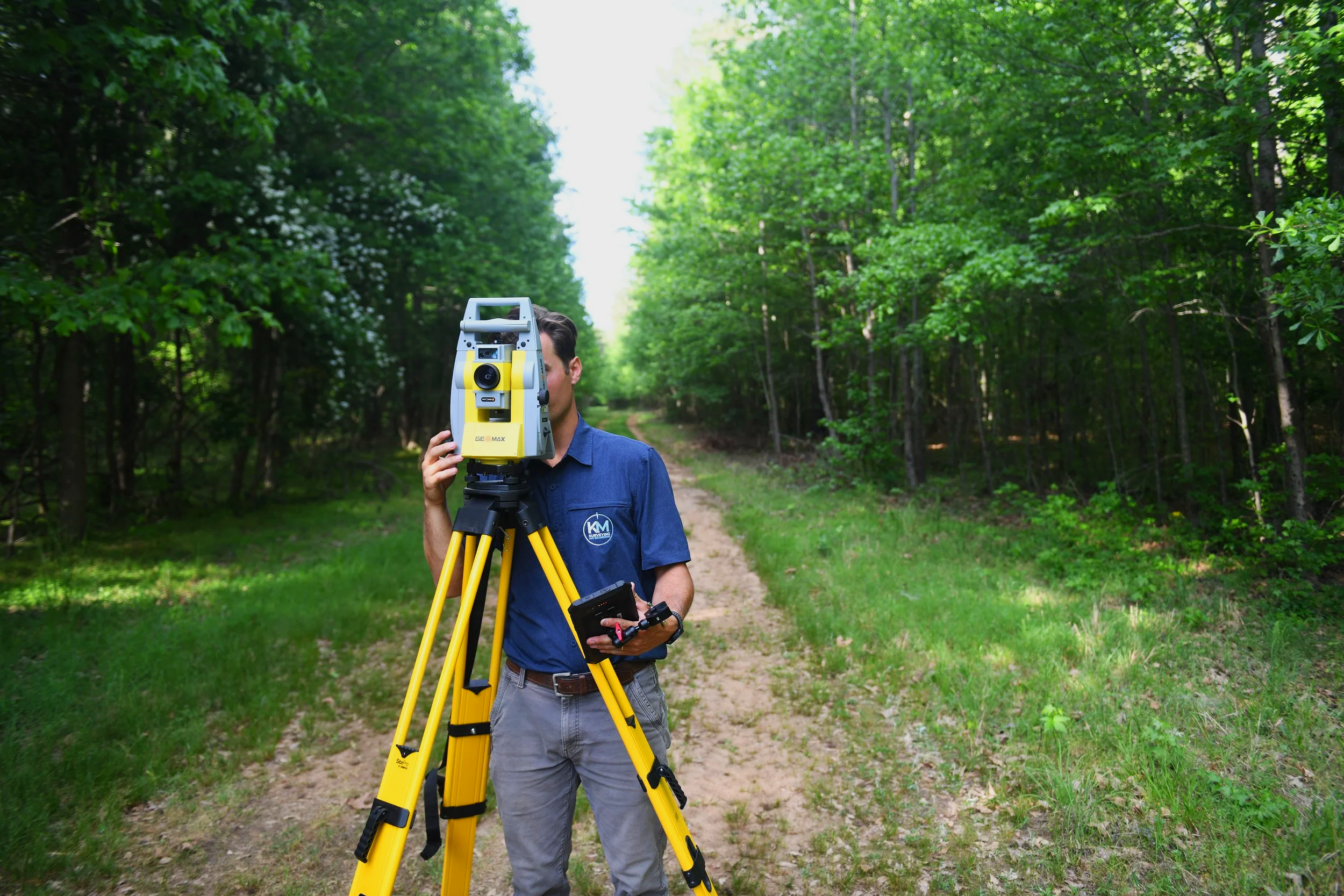 A man holding a surveying instrument on a tripod in a forested area with a dirt path and lush green trees.