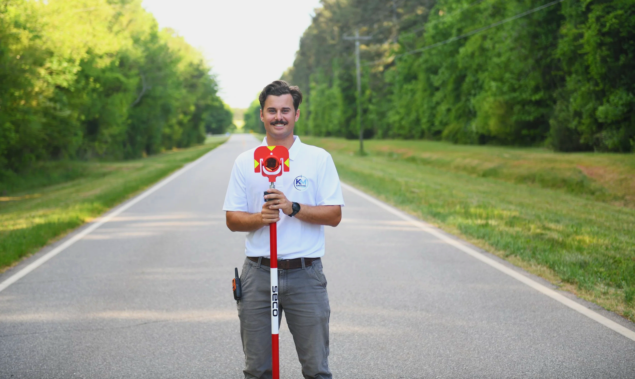 Man standing on a rural road with a surveying instrument, surrounded by lush green trees.