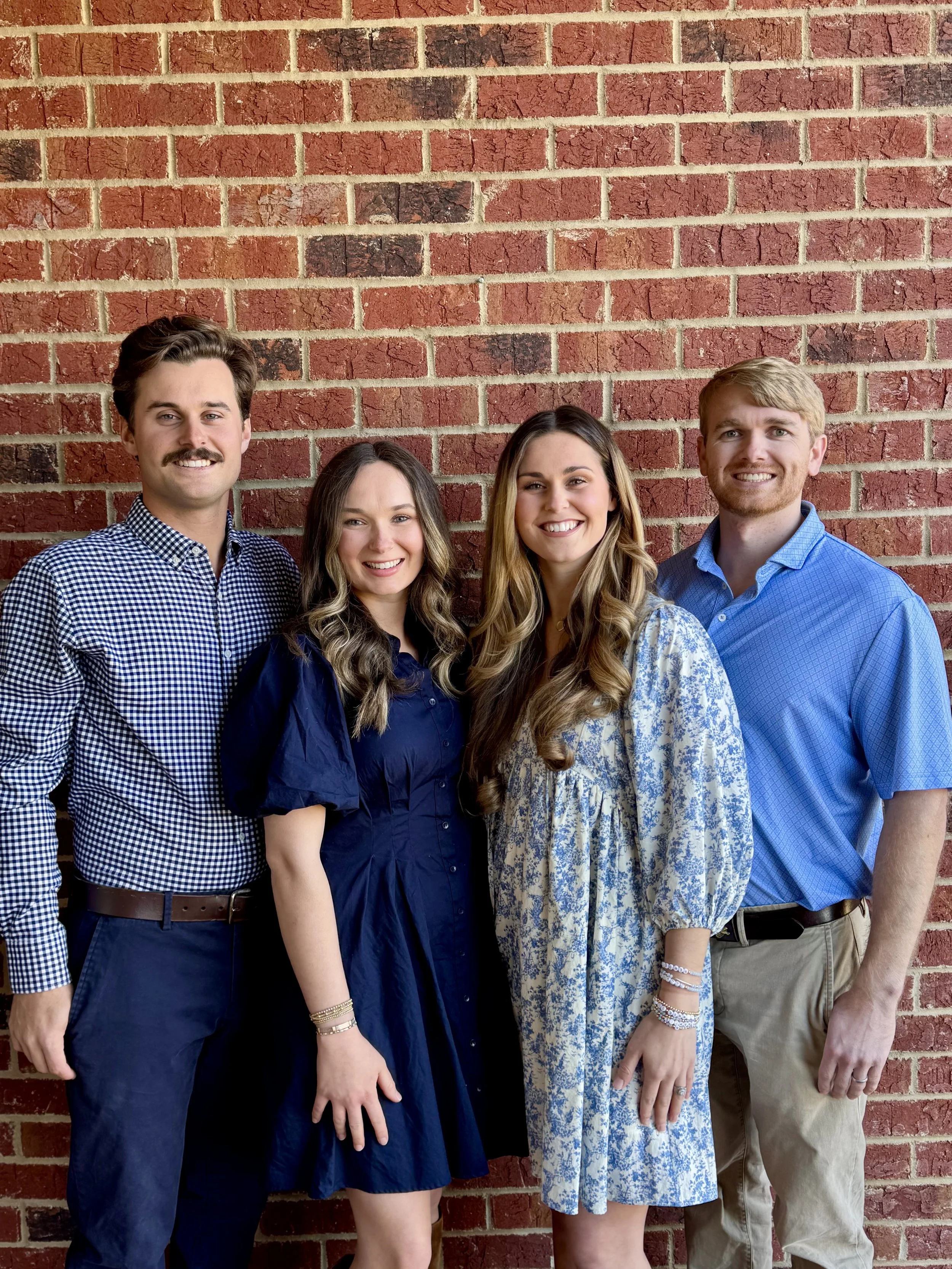 Four young adults standing in front of a red brick wall, smiling and posing for the photo.