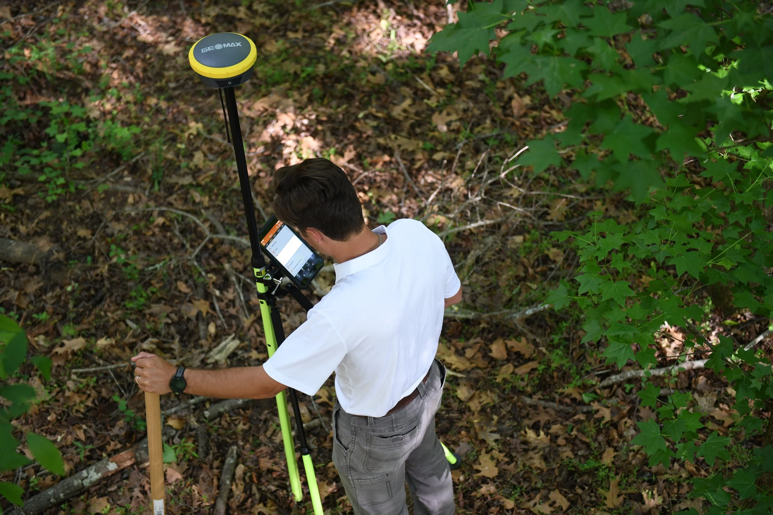 A man in a white shirt and gray pants using a GPS device in a forest, standing near a yellow and black Garmin GPS receiver mounted on a tripod, with green leaves and forest floor on the ground.
