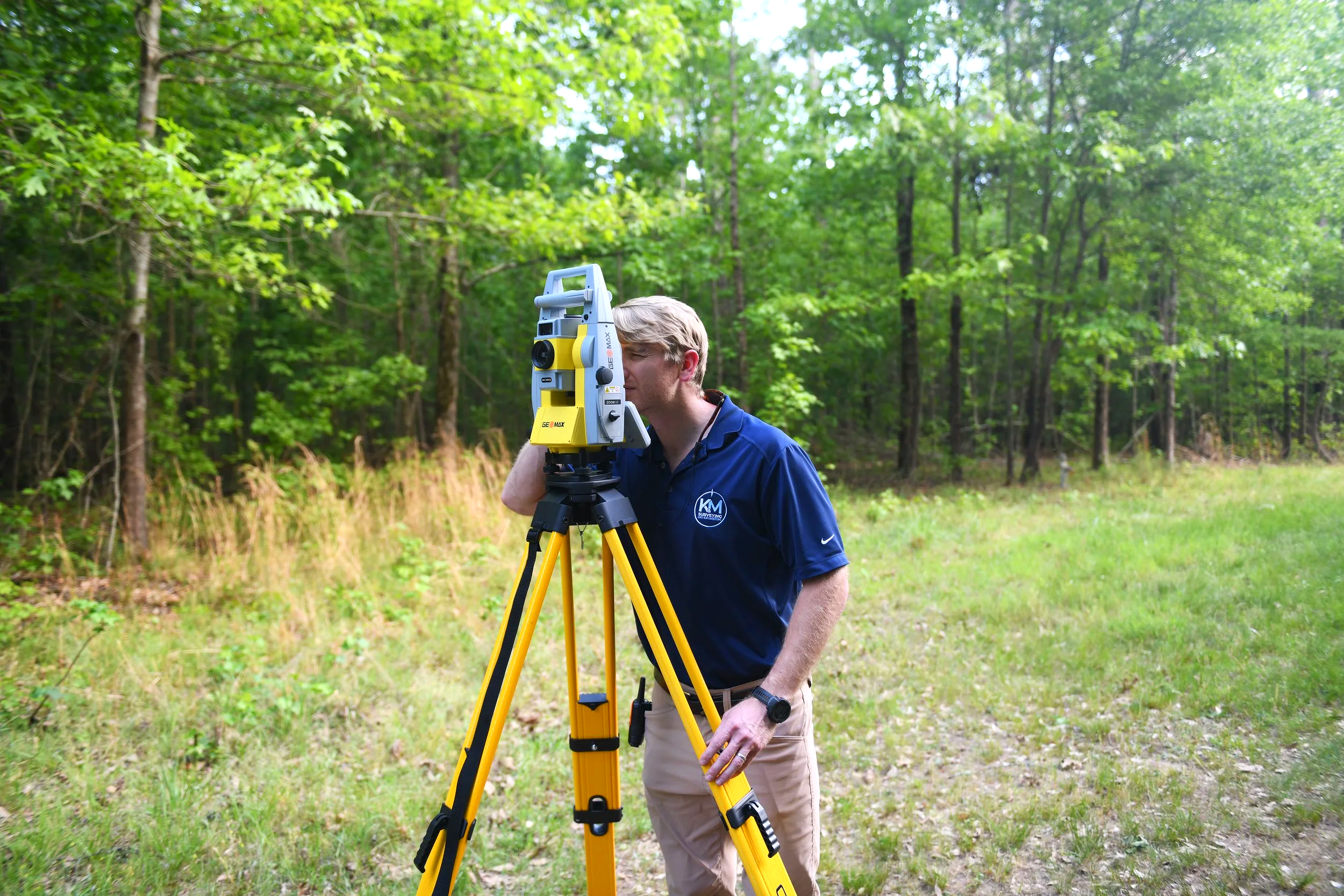 A surveyor in a blue shirt using a total station on a tripod in a wooded outdoor area.