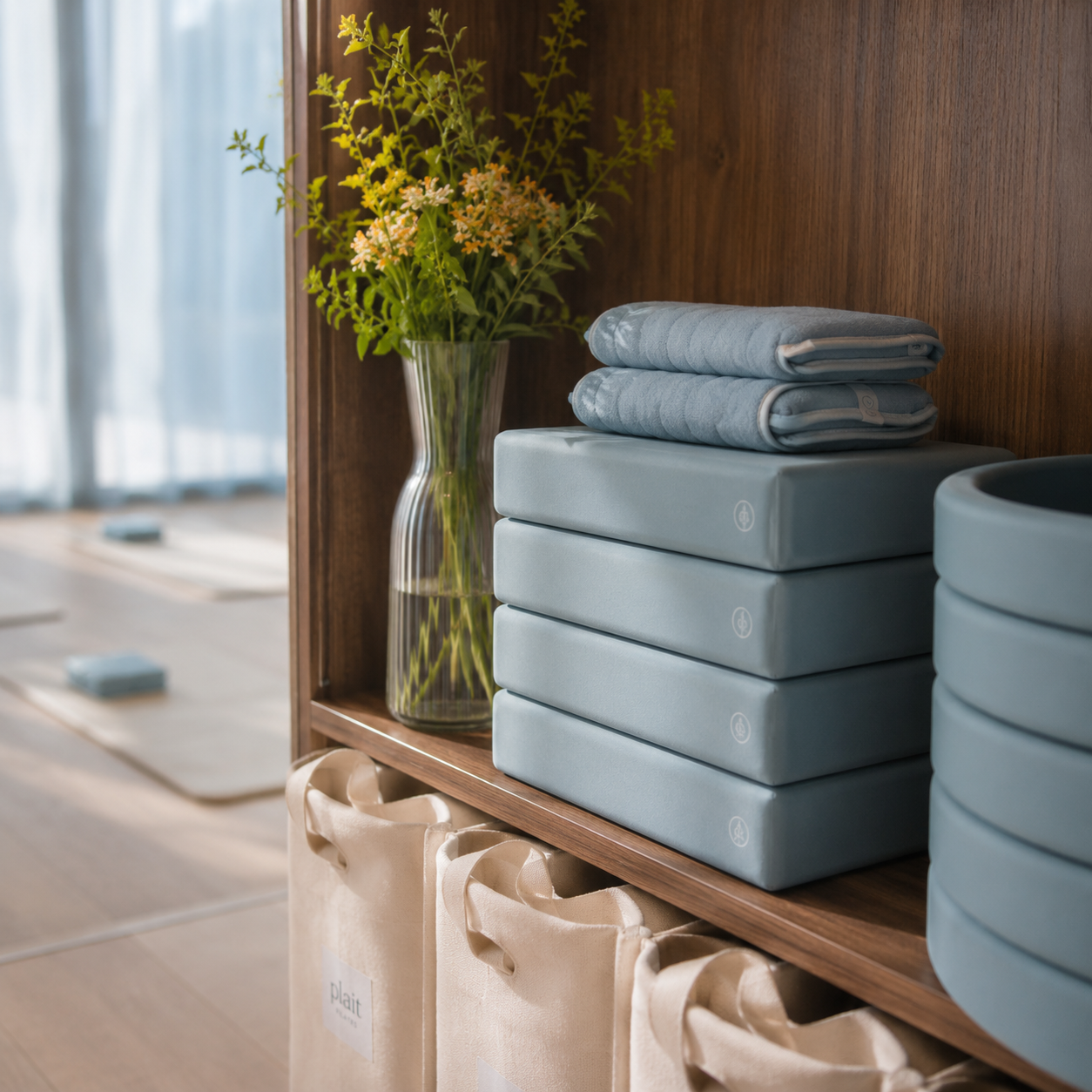 A wooden shelf with a tall glass vase containing green and orange flowers, neatly folded blue towels on stacked blue blocks, and beige tote bags hanging below.