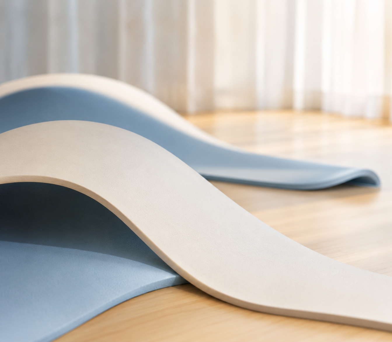 Close-up of two rolled pilates mats, one beige and one light blue, on a wooden floor in front of a window with sheer curtains
