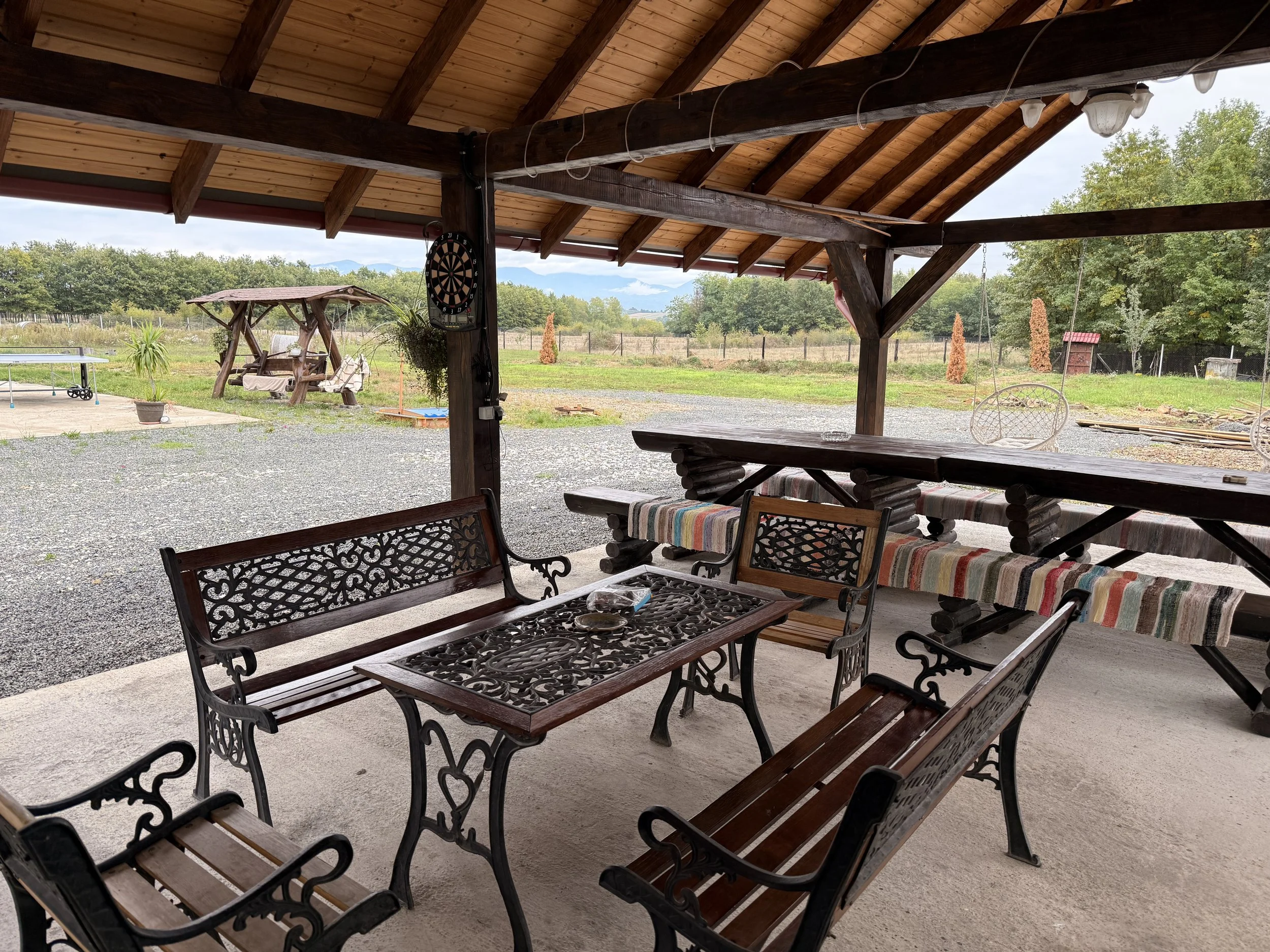 Outdoor covered patio with wooden furniture, including a table with four chairs, a long bench with striped cushion, and a table with a chessboard, overlooking a gravel yard with swings, a table tennis, and trees in the background.