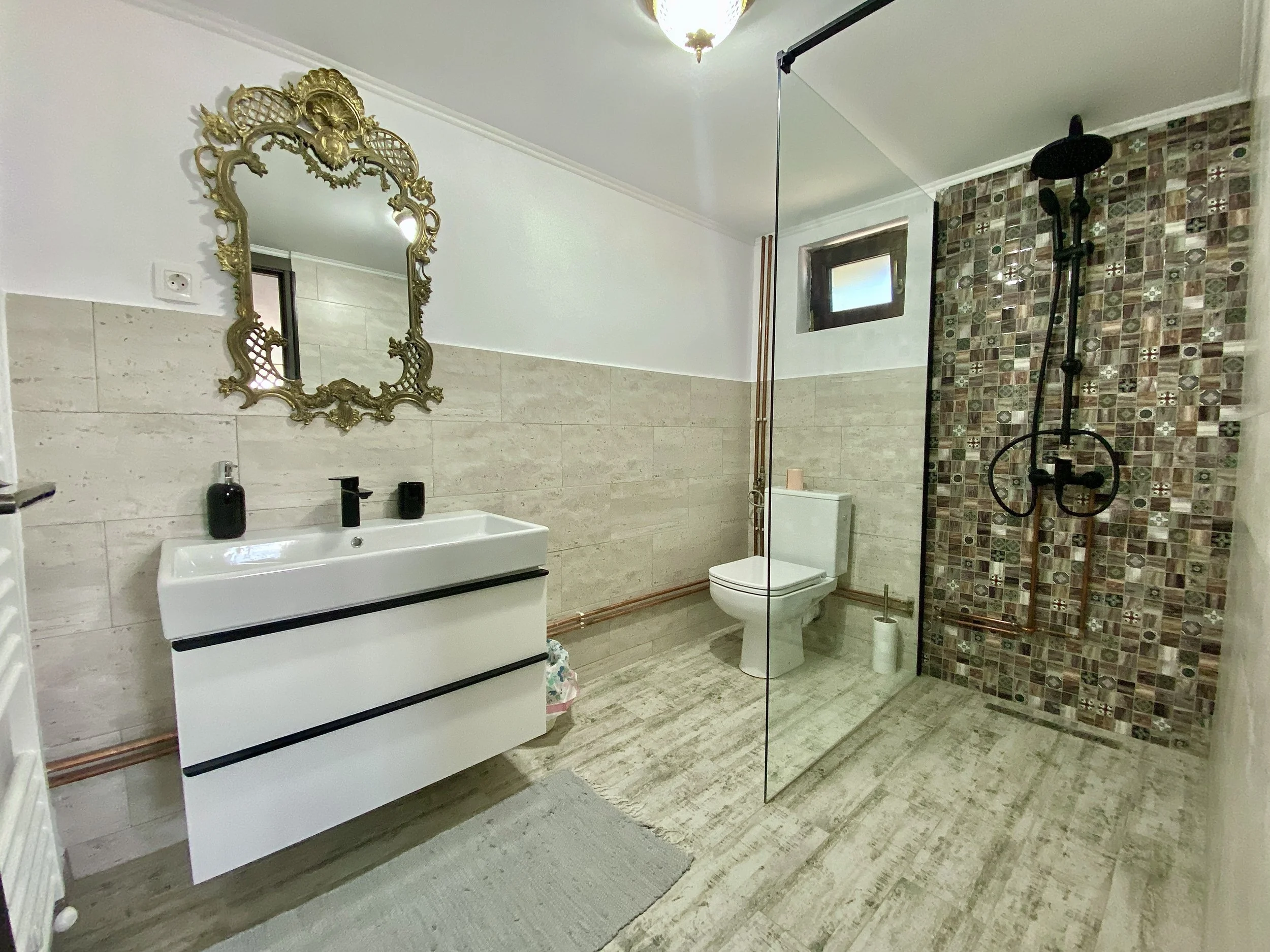 Bathroom with a white sink, gold ornate mirror, toilet, and walk-in shower with a glass partition and mosaic tile wall.