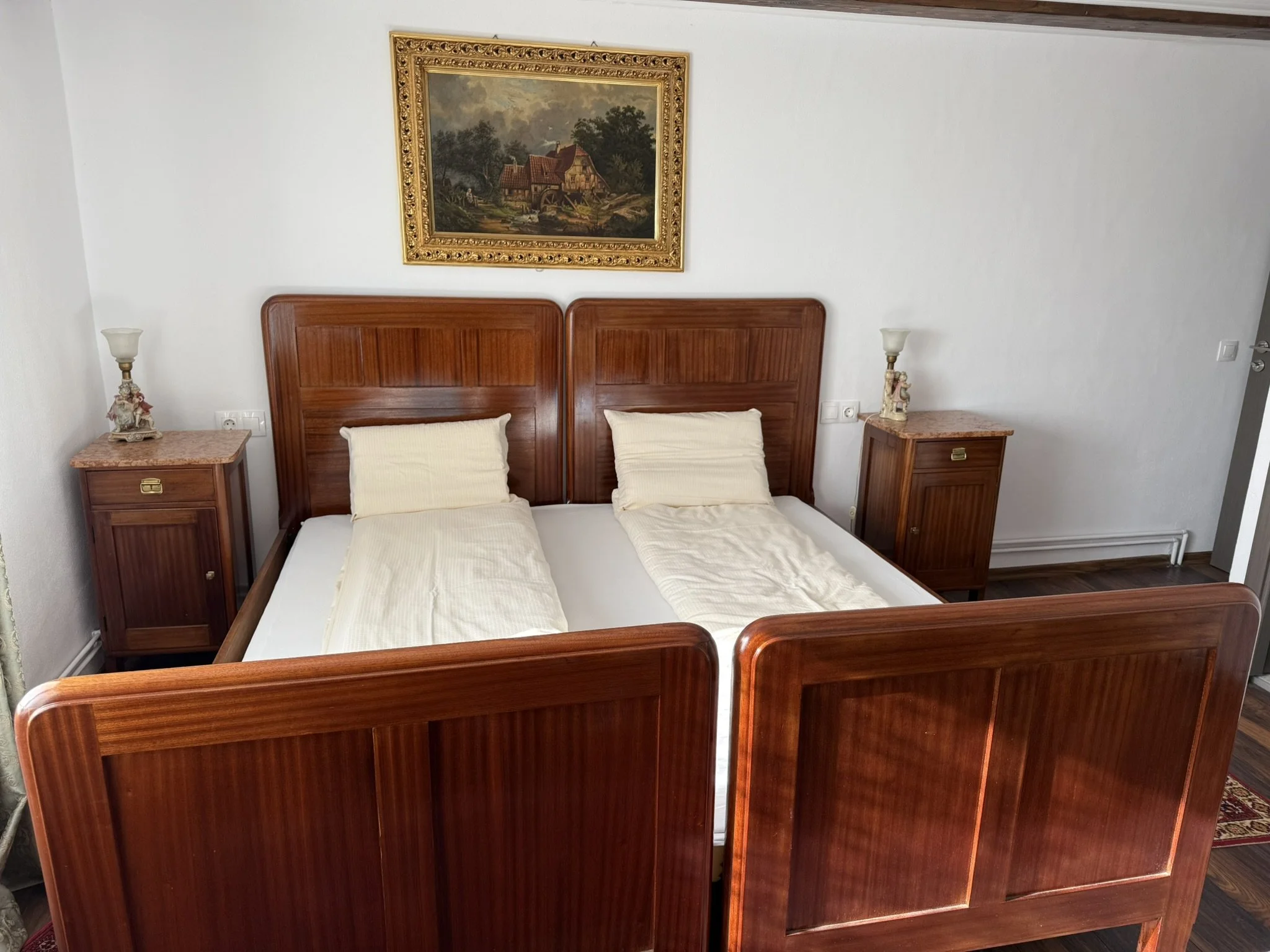 A bedroom with two single beds pushed together, each with a pillow and a cream-colored blanket. There are two wooden nightstands with lamps on either side of the beds. A framed painting of a countryside scene hangs above the headboard.