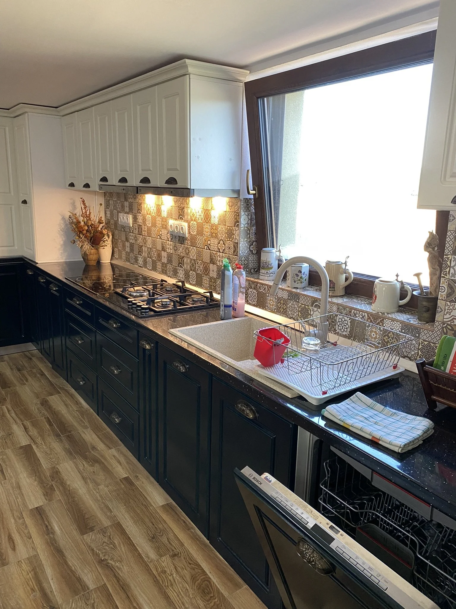 Kitchen with dark lower cabinets, cream upper cabinets, patterned backsplash, wooden flooring, and a large window above the sink.