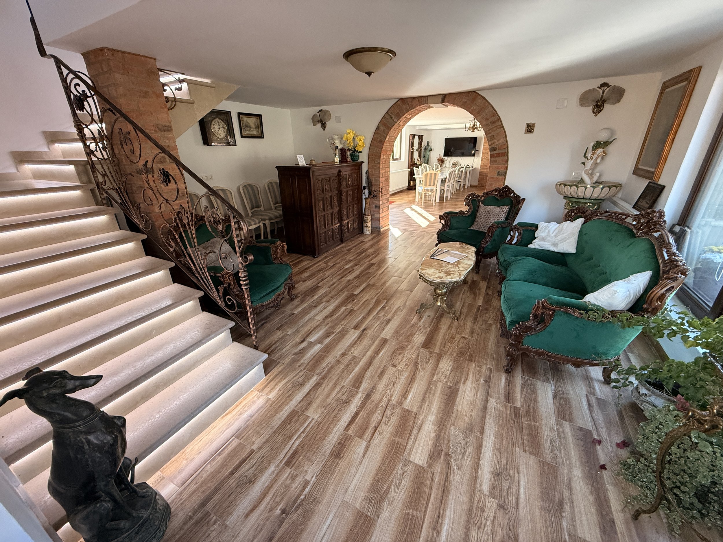 Living room with green antique furniture, wooden flooring, a staircase with ornate iron railing, and a brick archway leading to a dining area.