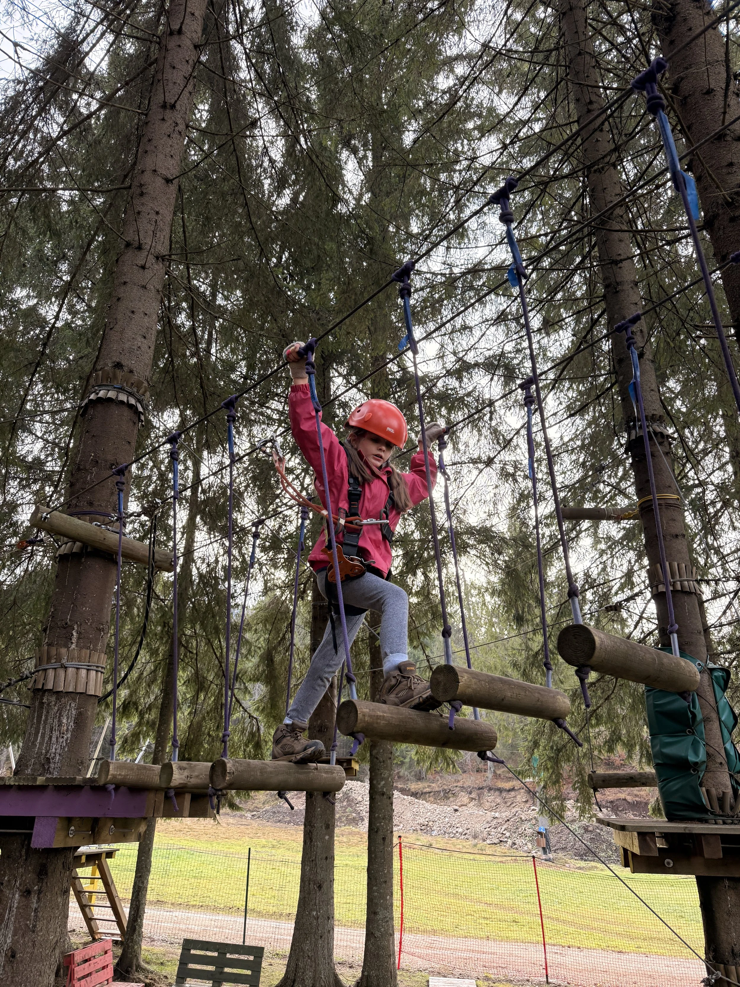 A young girl in a red helmet and pink jacket crossing a wooden bridge on a ropes course in a forested area.