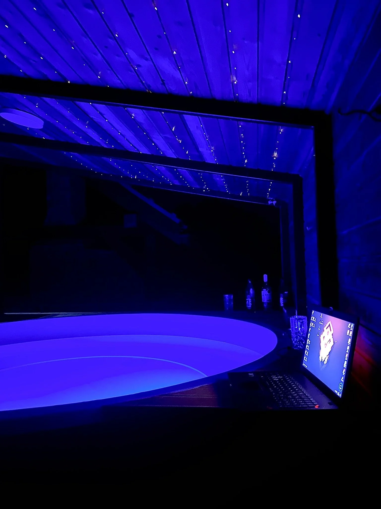 A room with a wooden ceiling decorated with small string lights, illuminated with blue lighting. There is a round illuminated DJ turntable, a laptop, and bottles placed on the shelf behind it. hot tub