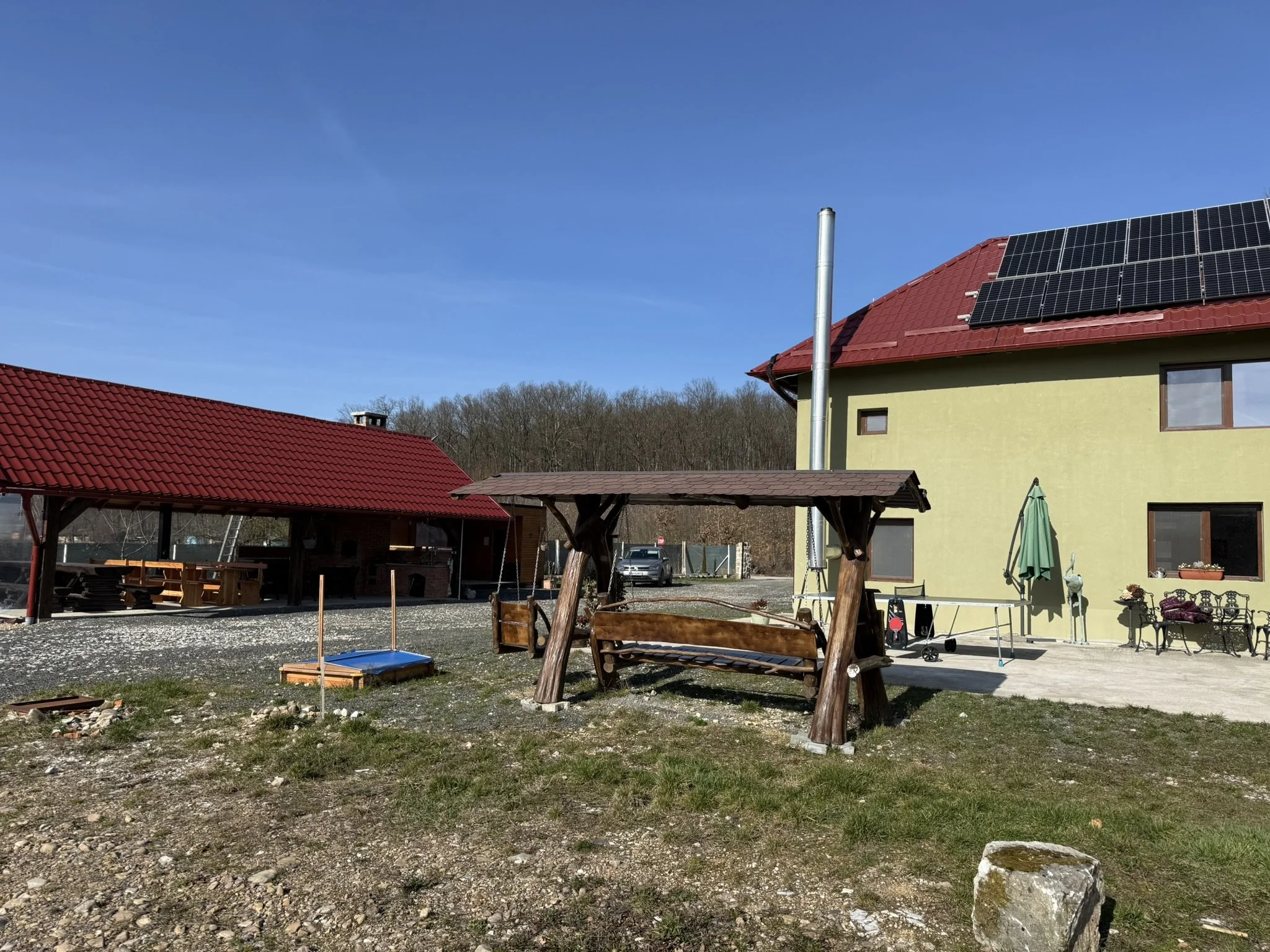 A yard with a swing, a covered seating area, a yellow house with solar panels, and a red-roofed building under a blue sky.