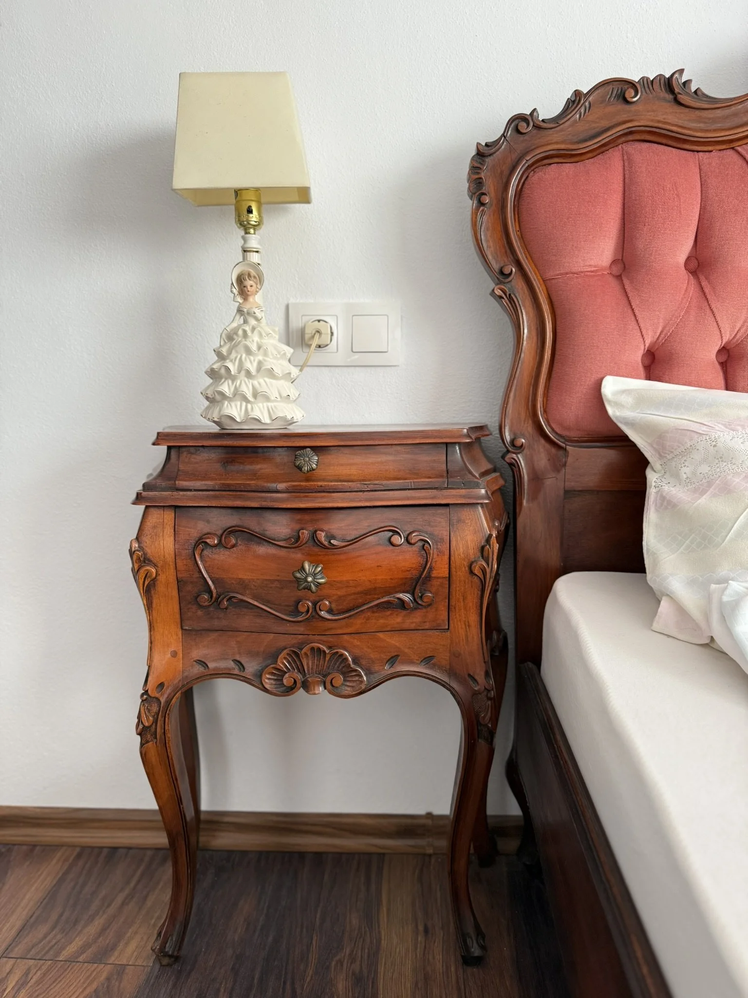 A vintage wooden nightstand with ornate carvings, next to a bed with a pink upholstered headboard, on a wooden floor.