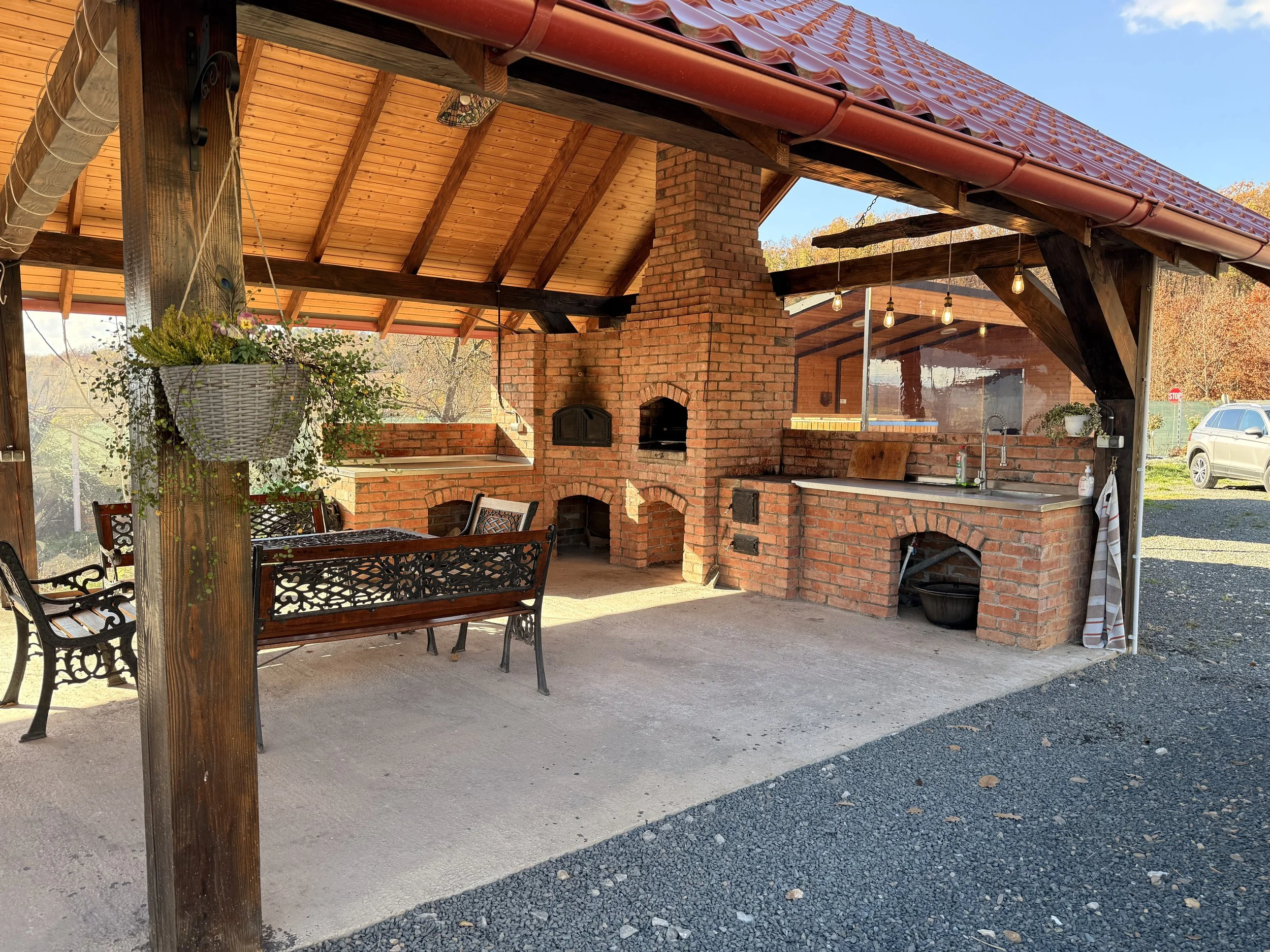 An outdoor covered patio with a brick fireplace, brick outdoor oven, and a kitchen sink area, with a wooden dining table and chairs, surrounded by trees with fall foliage.