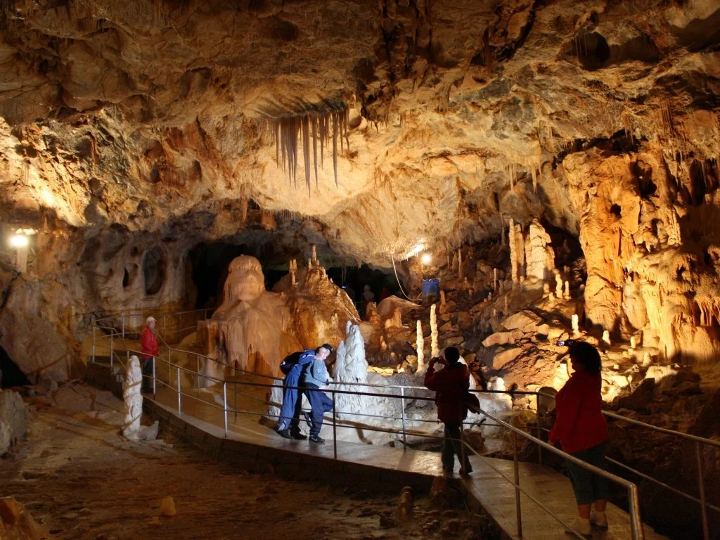 Visitors exploring a large, illuminated stalactite and stalagmite-filled cave with a wooden walkway for guided tours.