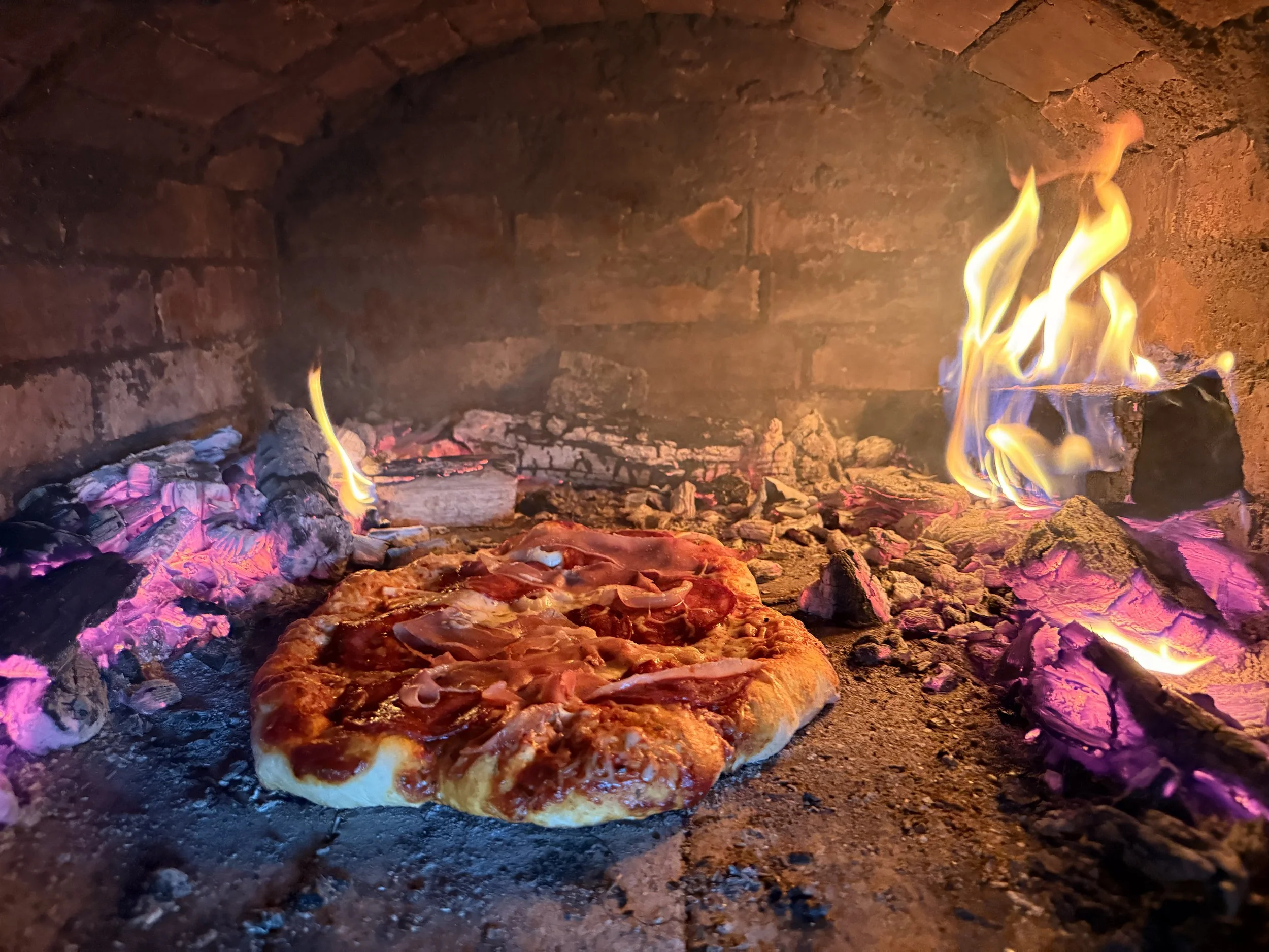 Pizza baking inside a brick oven with flames and glowing ashes.